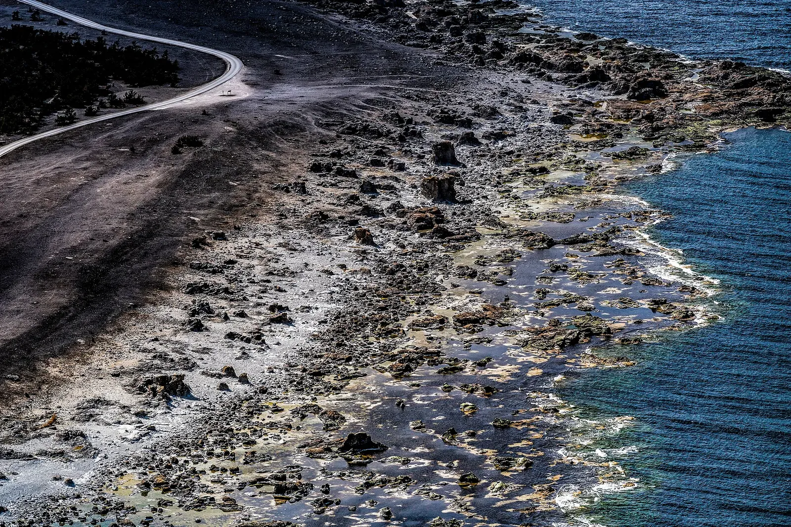 Aerial view of a flat grey limestone shore with rock pools and scattered stacks meeting dark blue water, a gravel road curving behind — Gotland & Fårö
