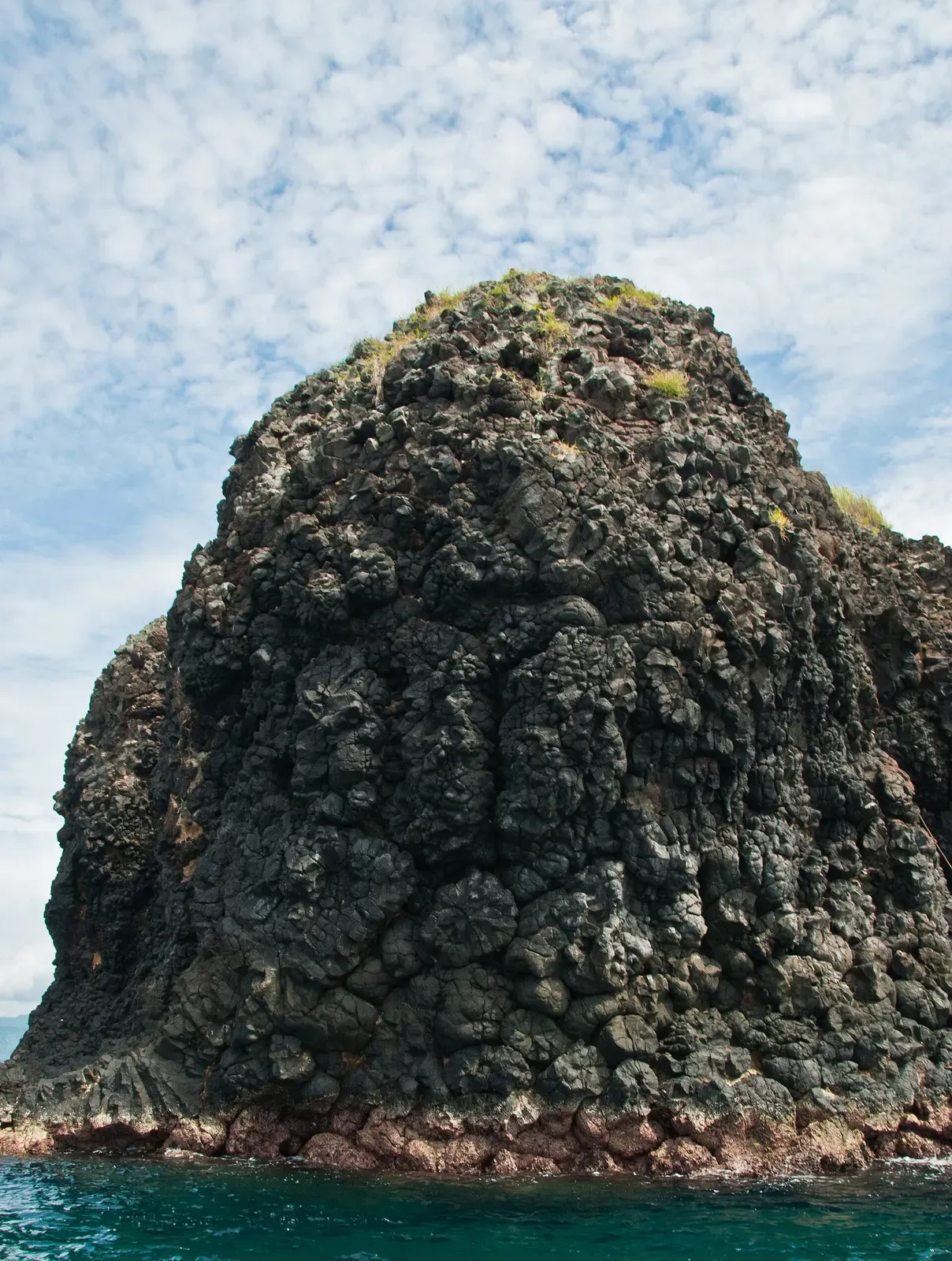 A tall rock islet of dark, rough volcanic rock rising from blue water — Gili Mimpang, Padangbai & Candidasa