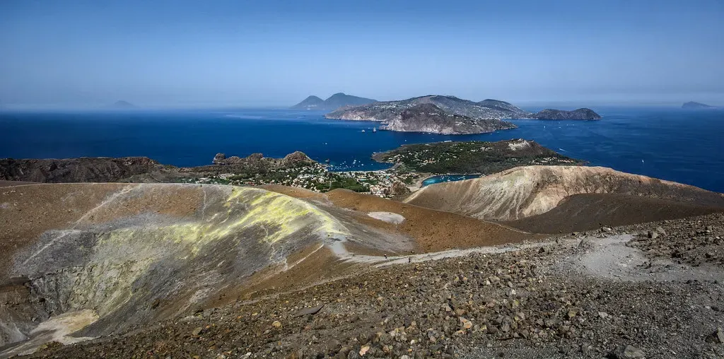 A yellow-streaked volcanic crater rim in the foreground, with a bay, a village and a rocky island across deep blue sea beyond — Aeolian Islands