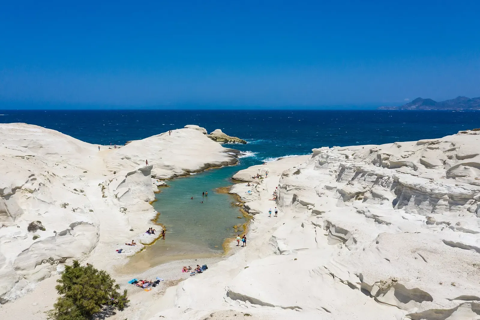Aerial view of white volcanic rock around a narrow turquoise inlet opening to dark blue sea, with a few swimmers and sunbathers — Milos & Kimolos