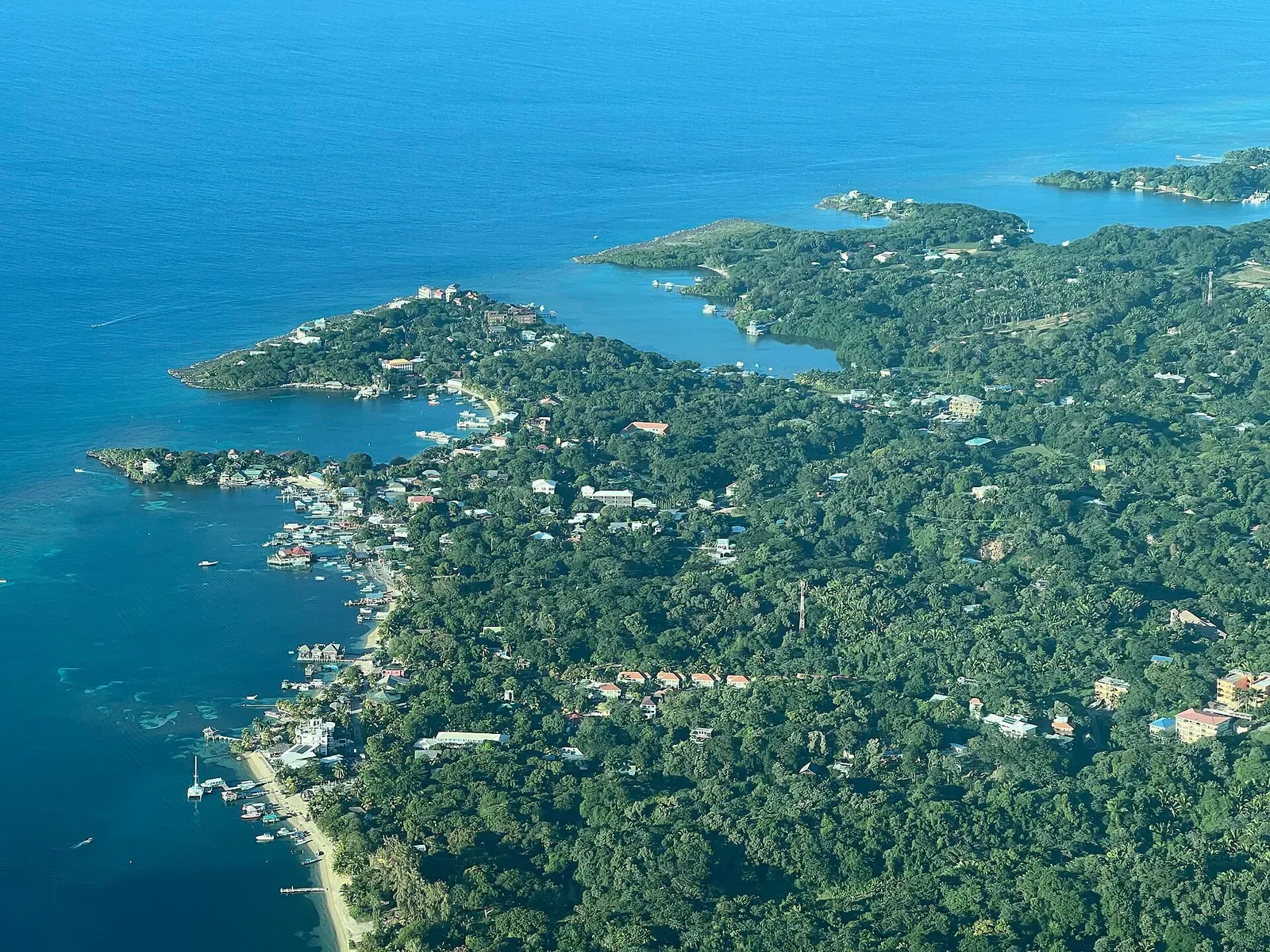 Aerial view of a forested coastline with small bays, docks and moored boats, and darker reef patches in the blue water offshore — West End Wall, Roatán