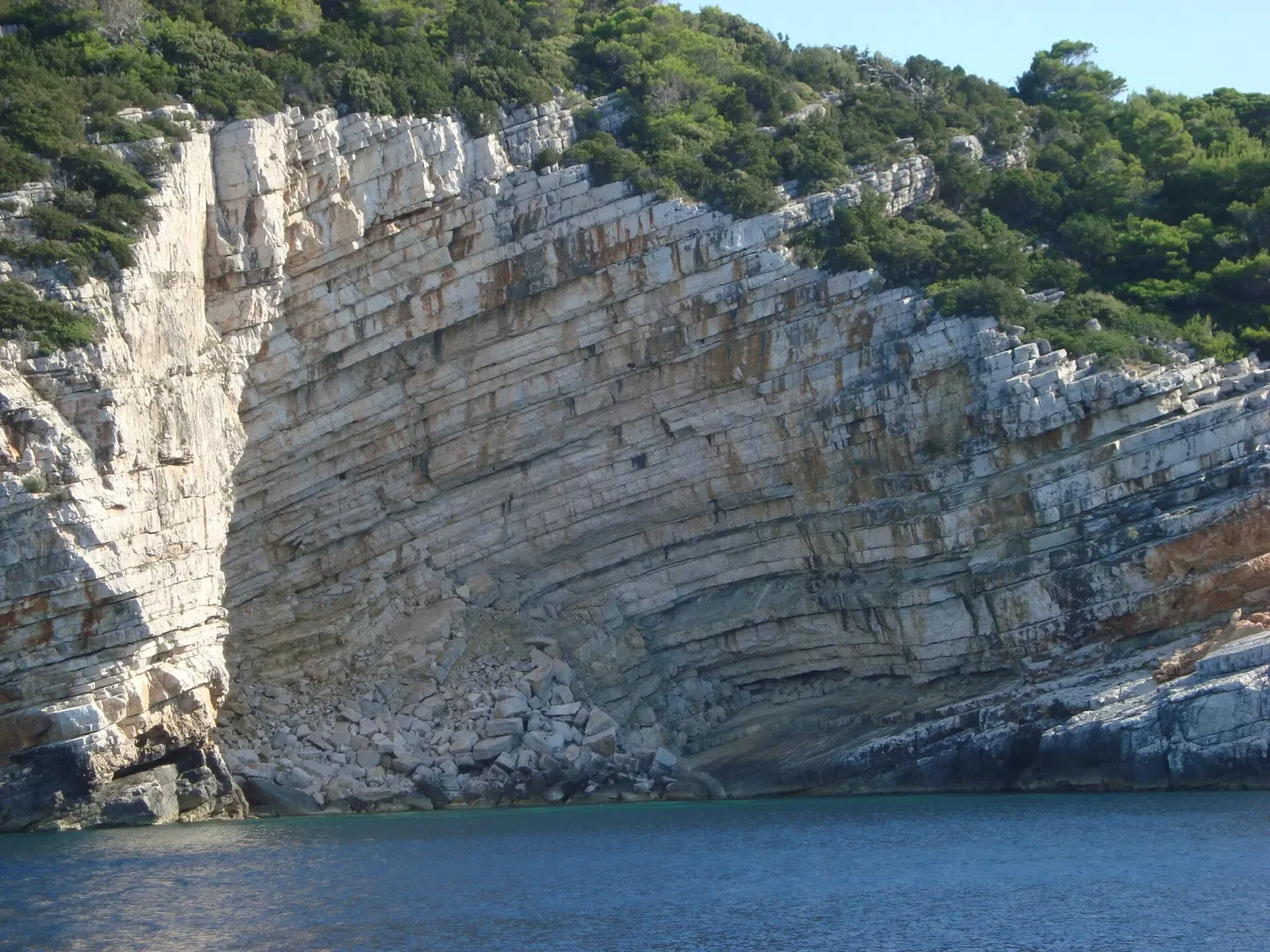 A pale layered limestone cliff curving down to a rubble slope at the waterline, with pine scrub above and calm blue sea below — Mljet National Park