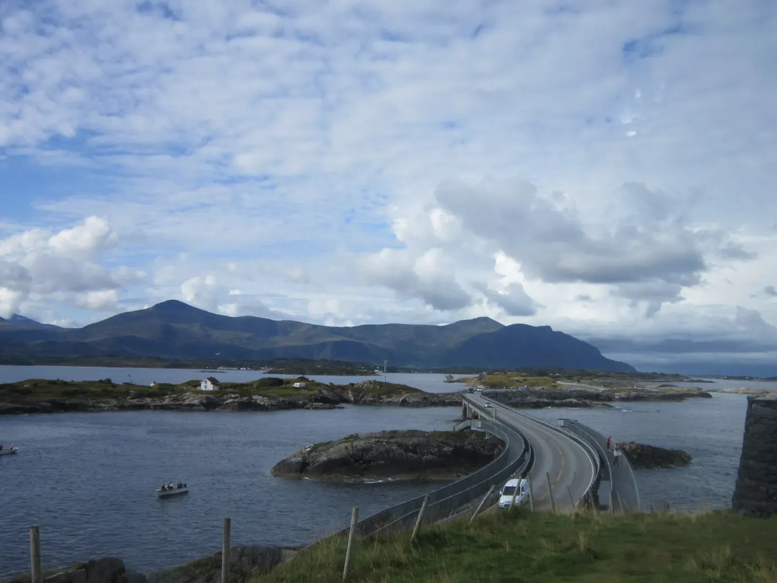 A road curving over low rocky islets between grey-blue water, with a small boat offshore and mountains under a cloudy sky — Kristiansund & the Atlantic Ocean Road.