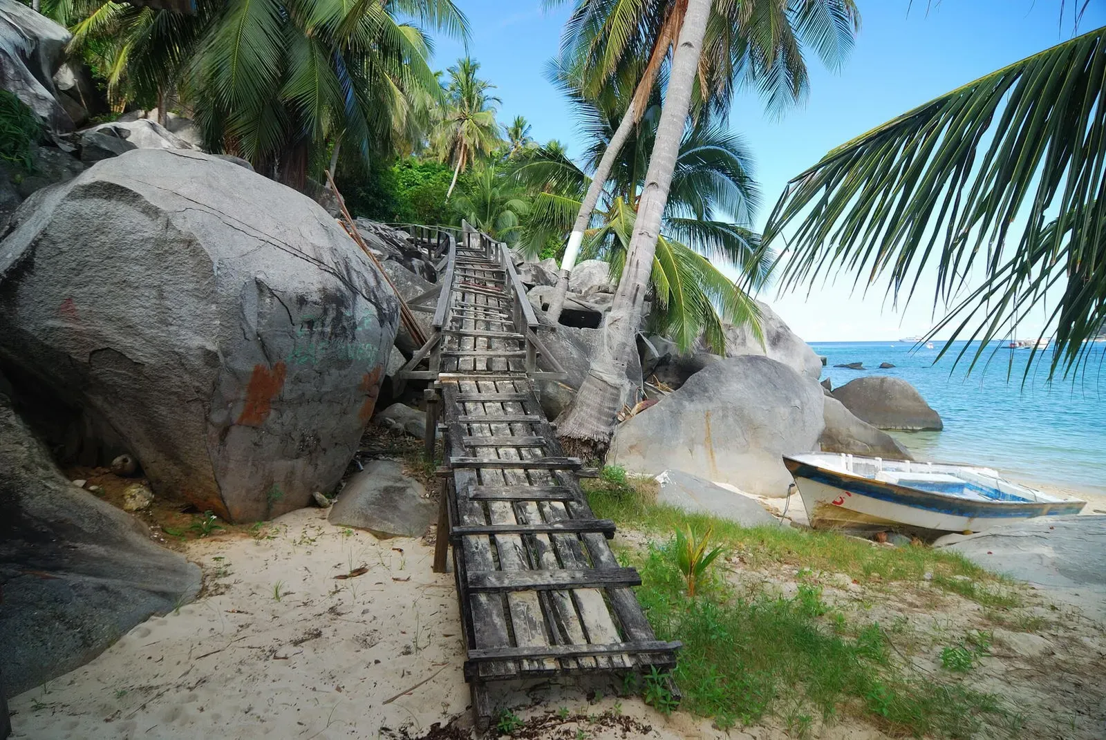 Wooden boardwalk climbing between large grey boulders and coconut palms, a small boat pulled up on the sand beside clear blue water — Pulau Aur & Pulau Dayang