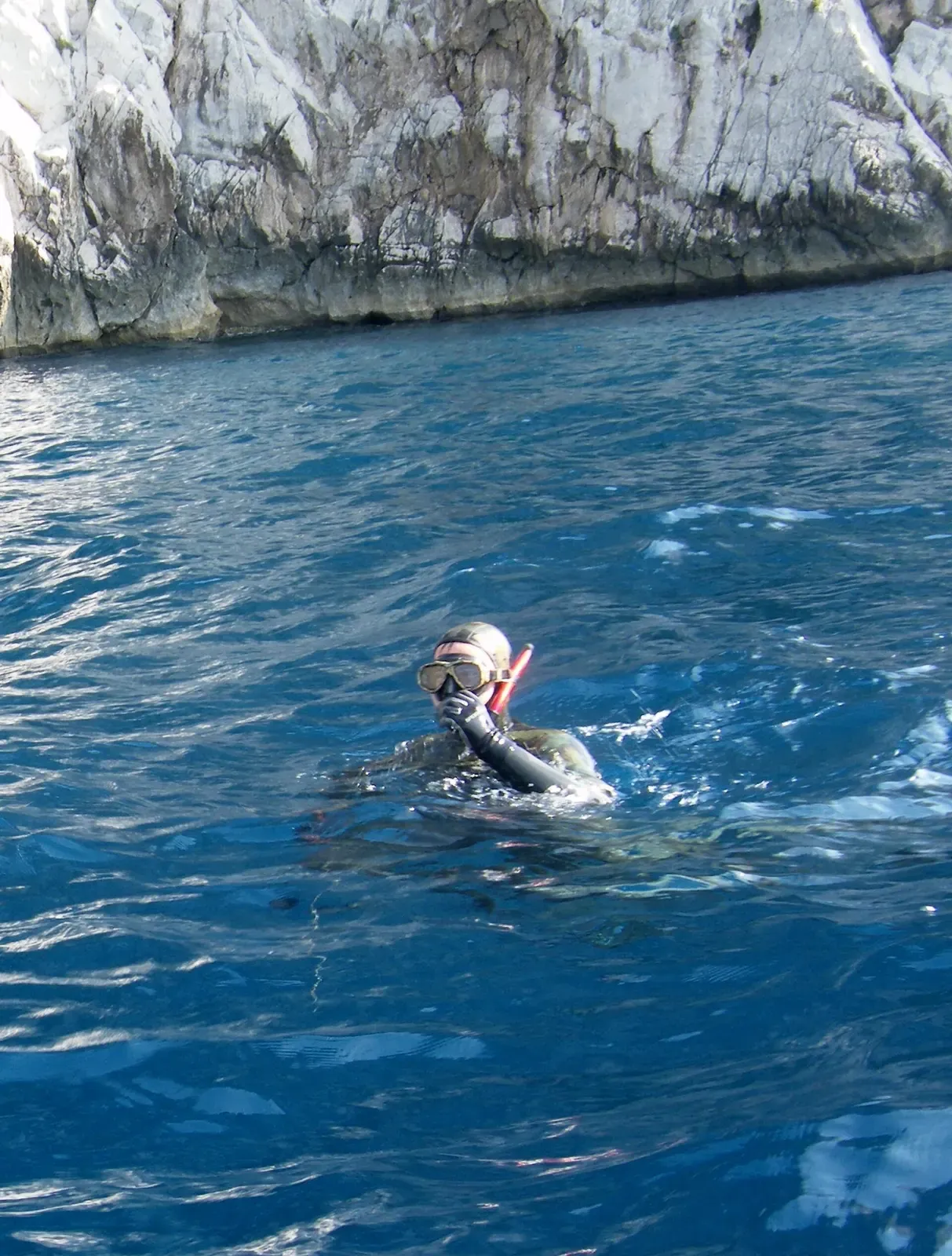 A person in a dark wetsuit, mask and red snorkel floating in blue water in front of a pale limestone cliff — Capri.