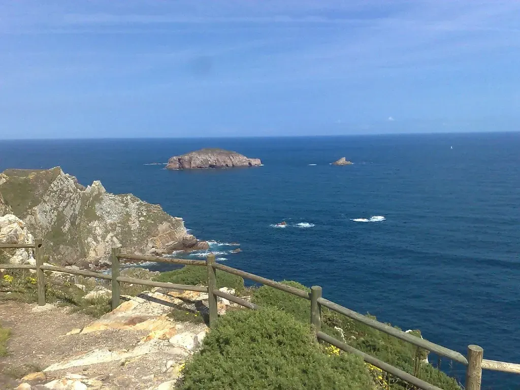 A wooden fence runs along a clifftop path above deep blue sea, with a rocky headland on the left and a low islet offshore — Gijón & Cabo Peñas