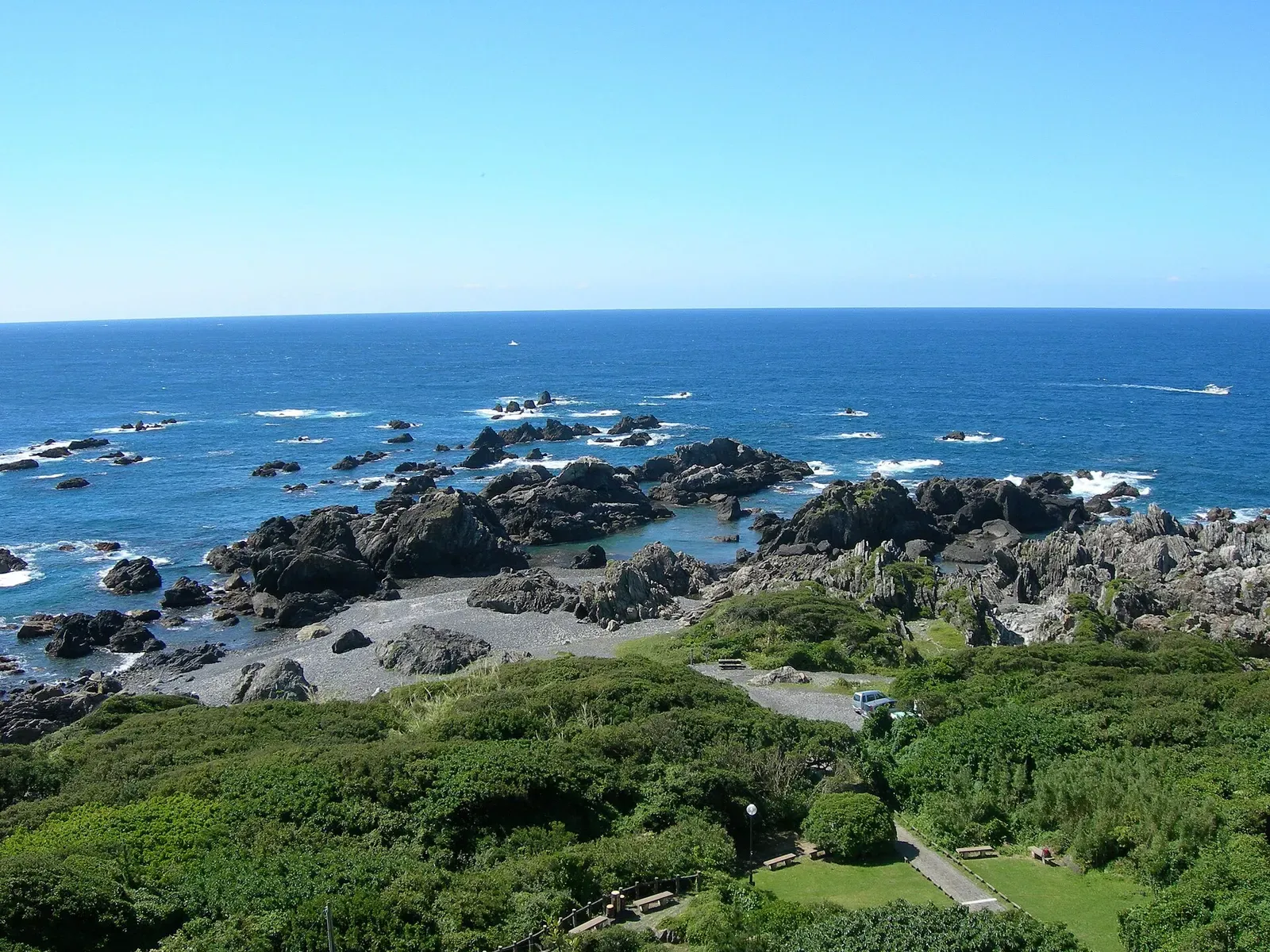 Dark rocky shoreline and scattered offshore rocks below green scrub, with deep blue open sea under a clear sky — Muroto, Kannoura & Shishikui