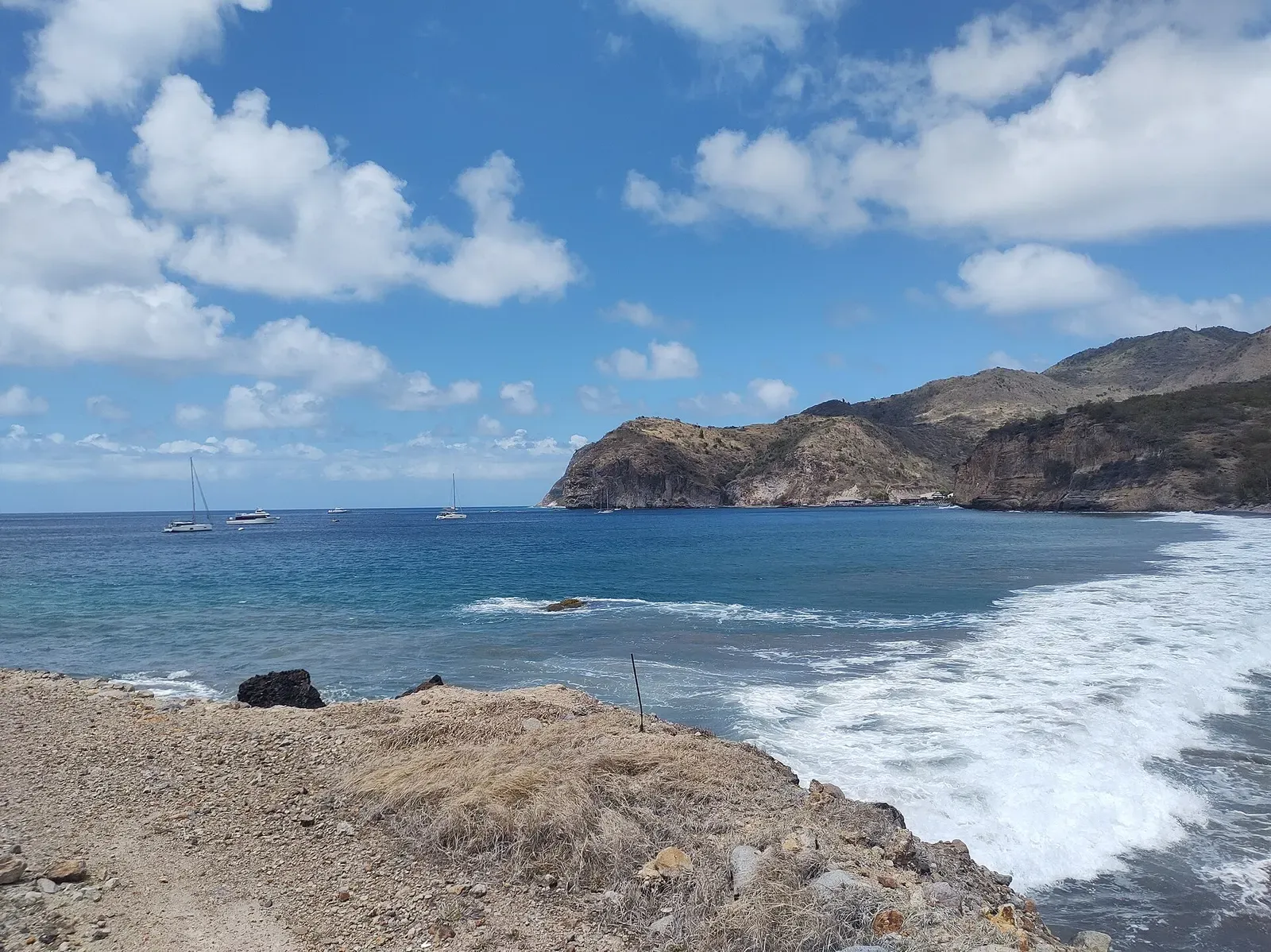 Rocky shoreline with white surf breaking below green headlands and sailboats anchored offshore under a blue sky — Montserrat
