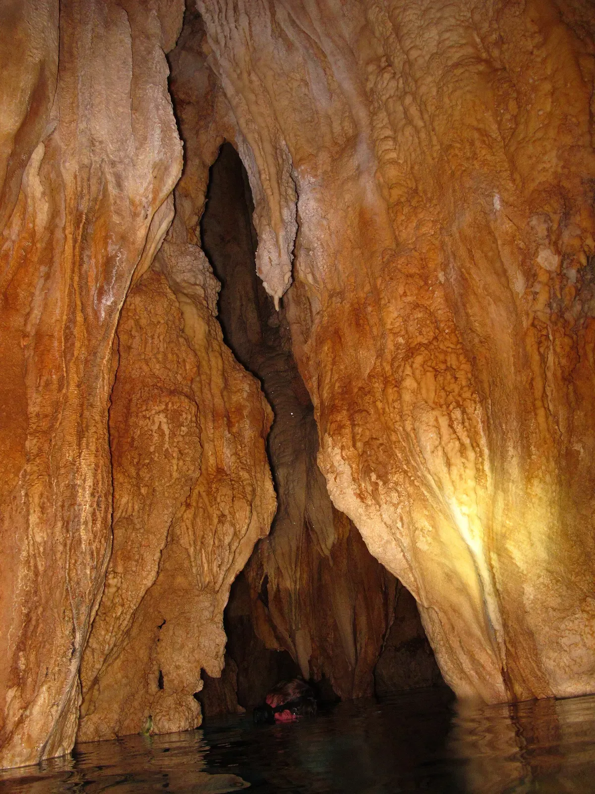 Orange-brown cave rock with hanging formations above dark still water inside a chamber — Chandelier Cave, Palau.