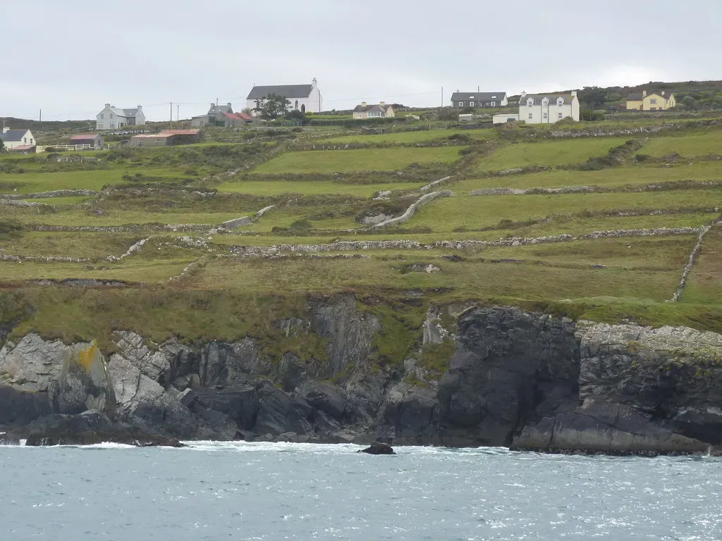 Dark rock cliffs rising from grey-green sea to stone-walled green fields and white houses on the hill above — Baltimore & Cape Clear Island