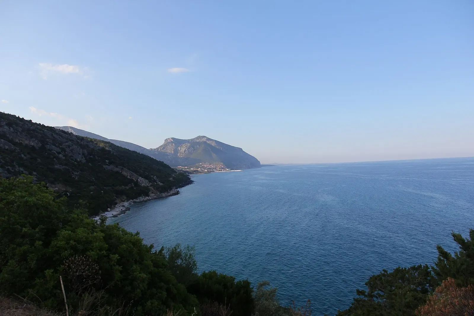 Calm blue sea below a wooded slope, with a rocky headland across the bay under a clear sky — Cala Gonone & the Gulf of Orosei