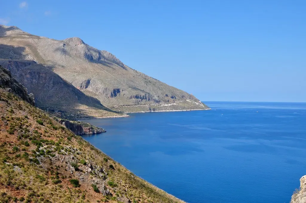 Steep scrub-covered mountainside dropping into deep blue sea, with a headland and a coastal track in the distance under a clear sky — San Vito Lo Capo & Scopello
