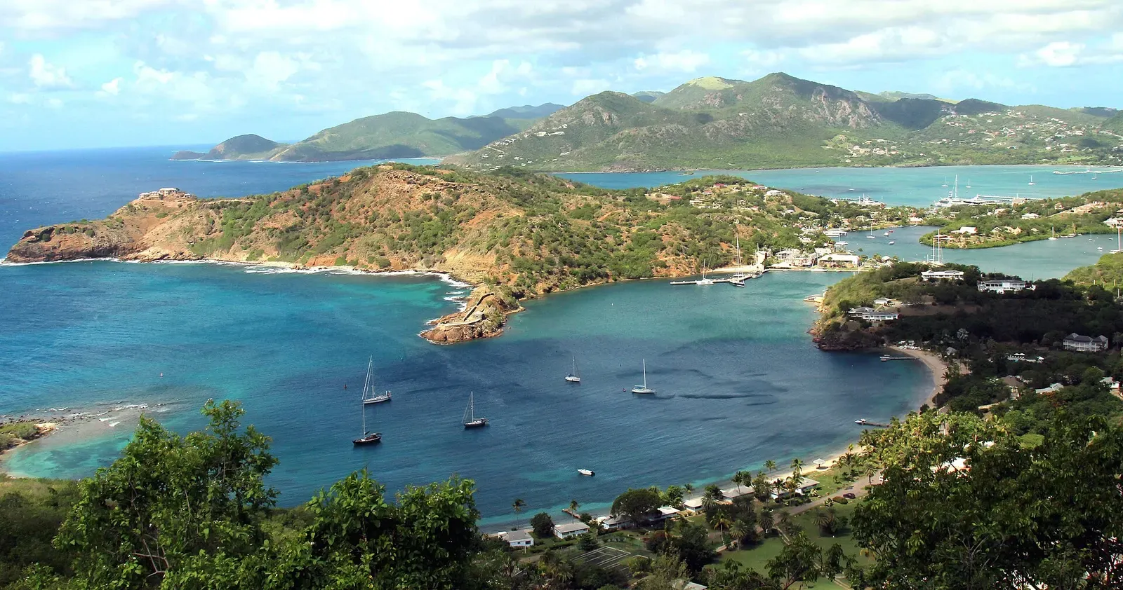 Aerial view over a turquoise bay with anchored sailboats, a scrub-covered headland and green hills beyond — Antigua