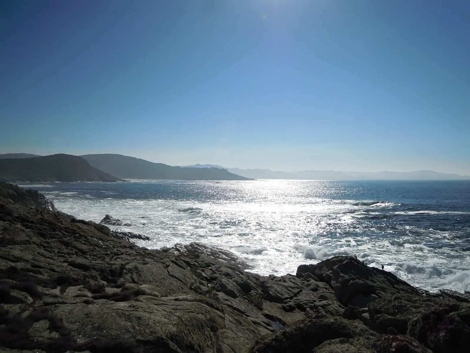 Dark granite rocks in the foreground, white surf breaking along the shore and headlands fading into haze under a clear blue sky — Costa da Morte