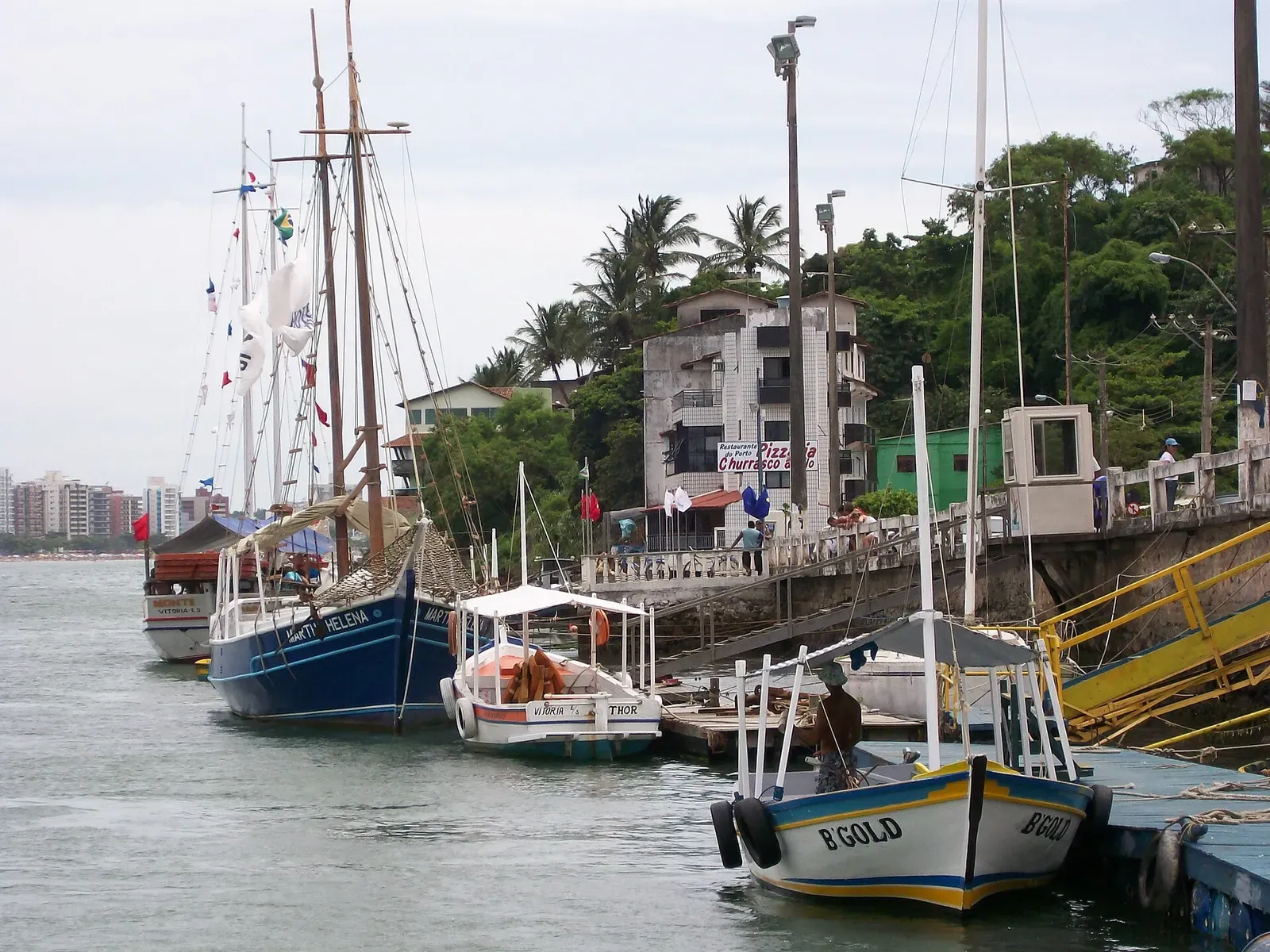 Two masted schooners and small boats moored along a quay, with palms and buildings on the shore behind — Guarapari.