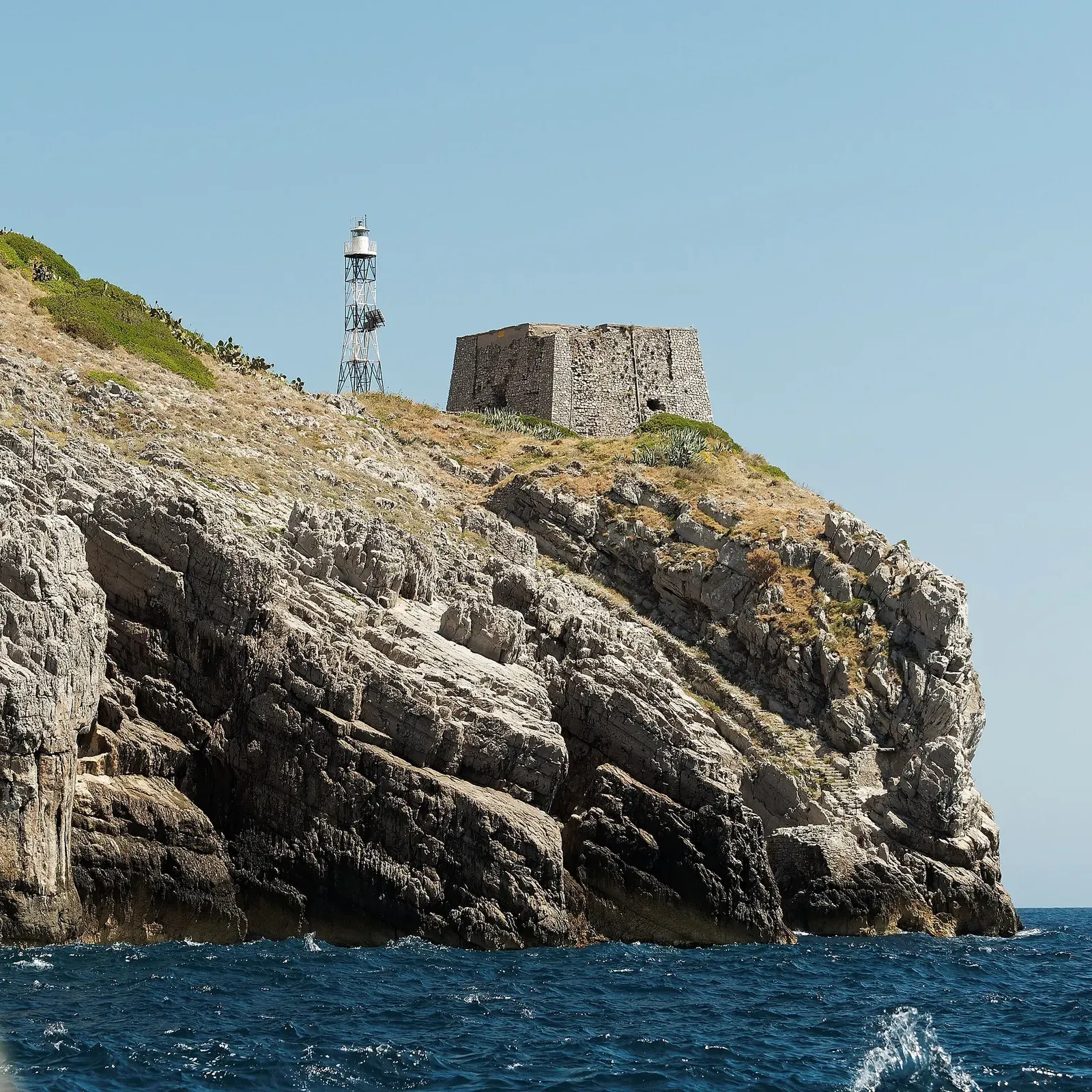 A steel lattice light tower and a square stone watchtower stand on a craggy limestone headland above dark blue water — Punta Campanella & the Sorrento Coast
