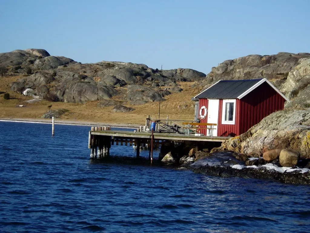 A red wooden boathouse with a small jetty on bare granite rock, beside deep blue water, with rounded grey rock hills behind — Gothenburg Archipelago