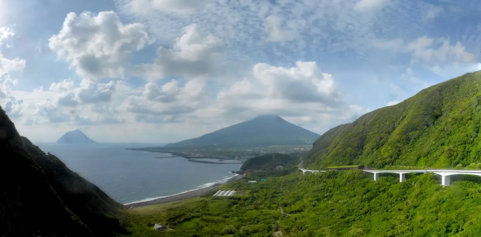 Green slopes and a road viaduct above a curved bay, with a cone-shaped volcano and a small steep island offshore under scattered cloud — Hachijojima