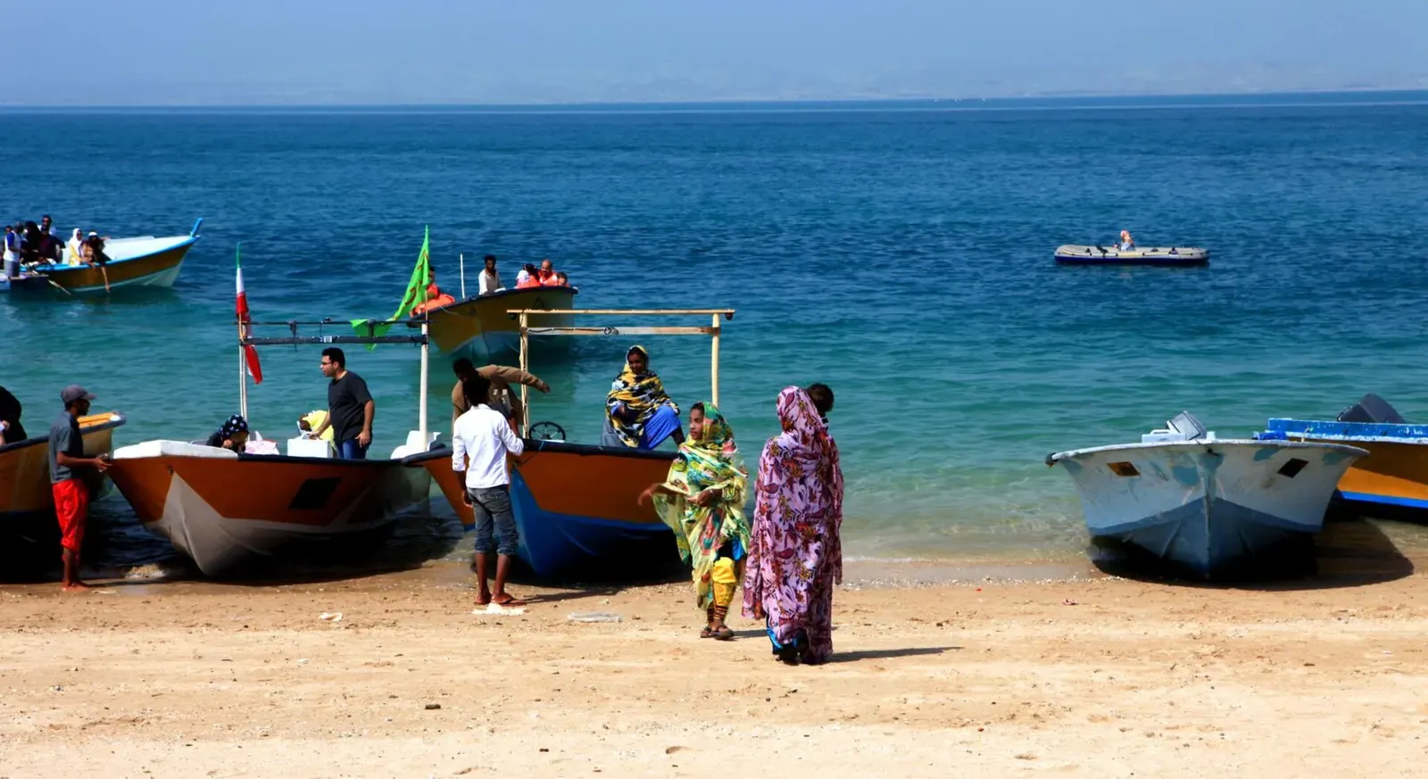 Painted wooden boats pulled up on a sandy beach beside clear blue water, with a few people on the sand and passengers in orange life jackets aboard — Qeshm & Hengam Island.