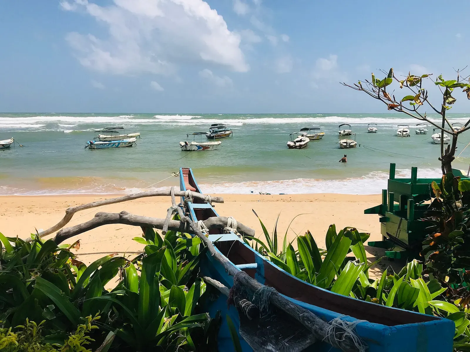 A blue wooden outrigger canoe among green plants on a sandy beach, with small motor boats moored in the bay beyond — Hikkaduwa.