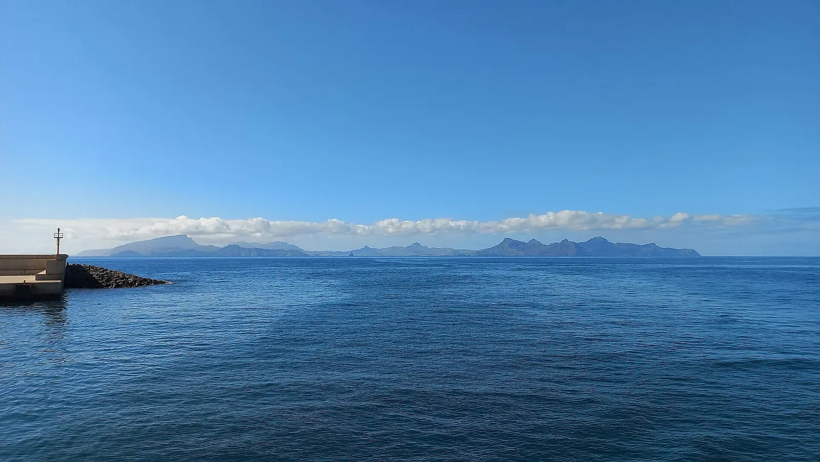 Calm blue sea under a clear sky, a harbour breakwater at left and a mountainous island on the horizon — Santo Antão