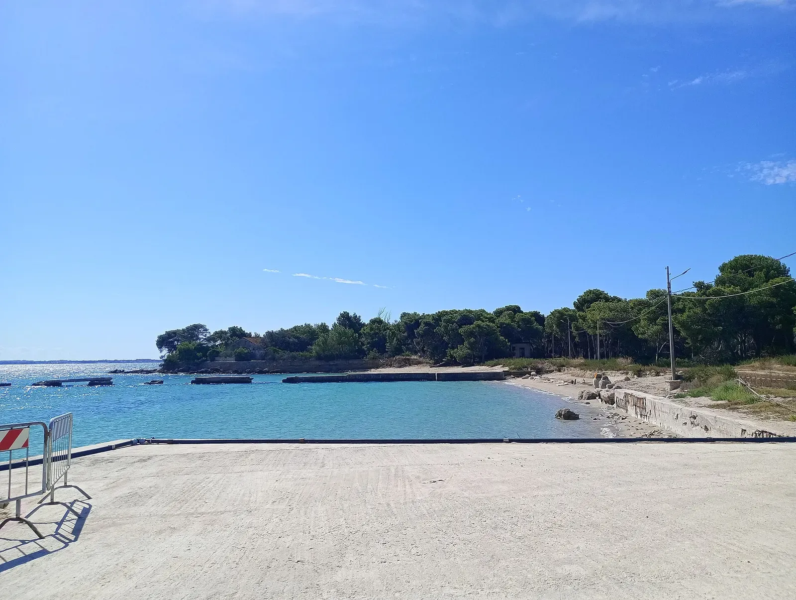 A pale concrete quay in front of a turquoise bay, an empty sandy beach and a pine-covered shoreline under a clear blue sky — Taranto & the Gulf of Taranto