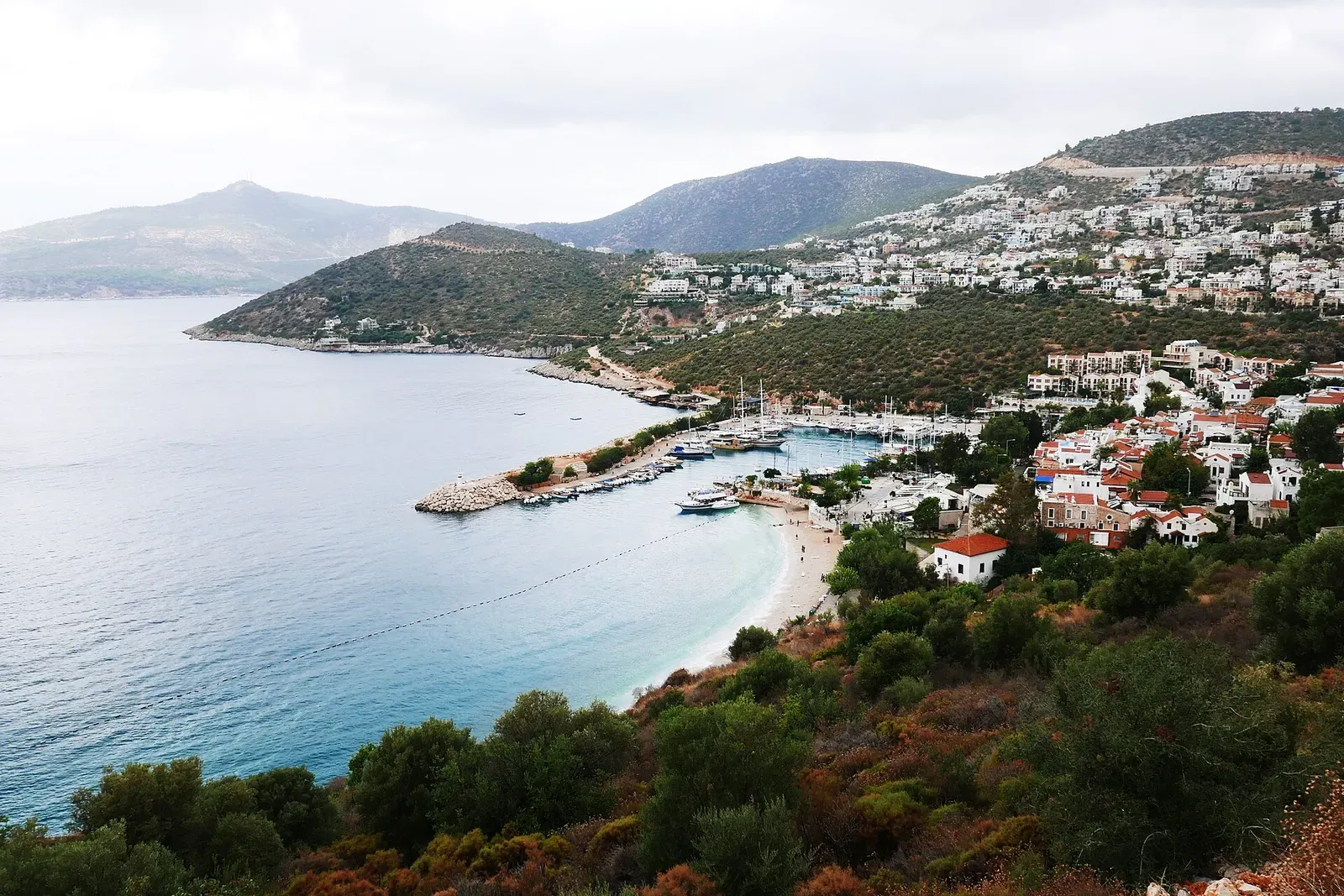 Kalkan Bay from a scrub-covered hillside: a curved pale beach, a harbour breakwater with moored boats, and white villas on green slopes