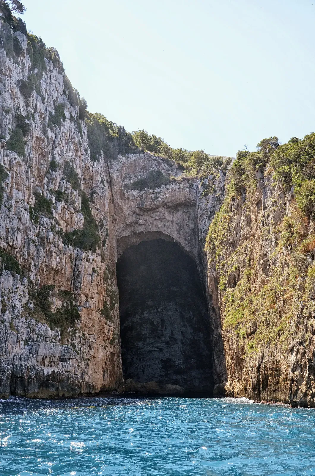 A tall dark cave mouth at the foot of sheer grey limestone cliffs topped with green scrub, above blue water — Haxhi Ali Cave, Vlora, Karaburun & Sazan