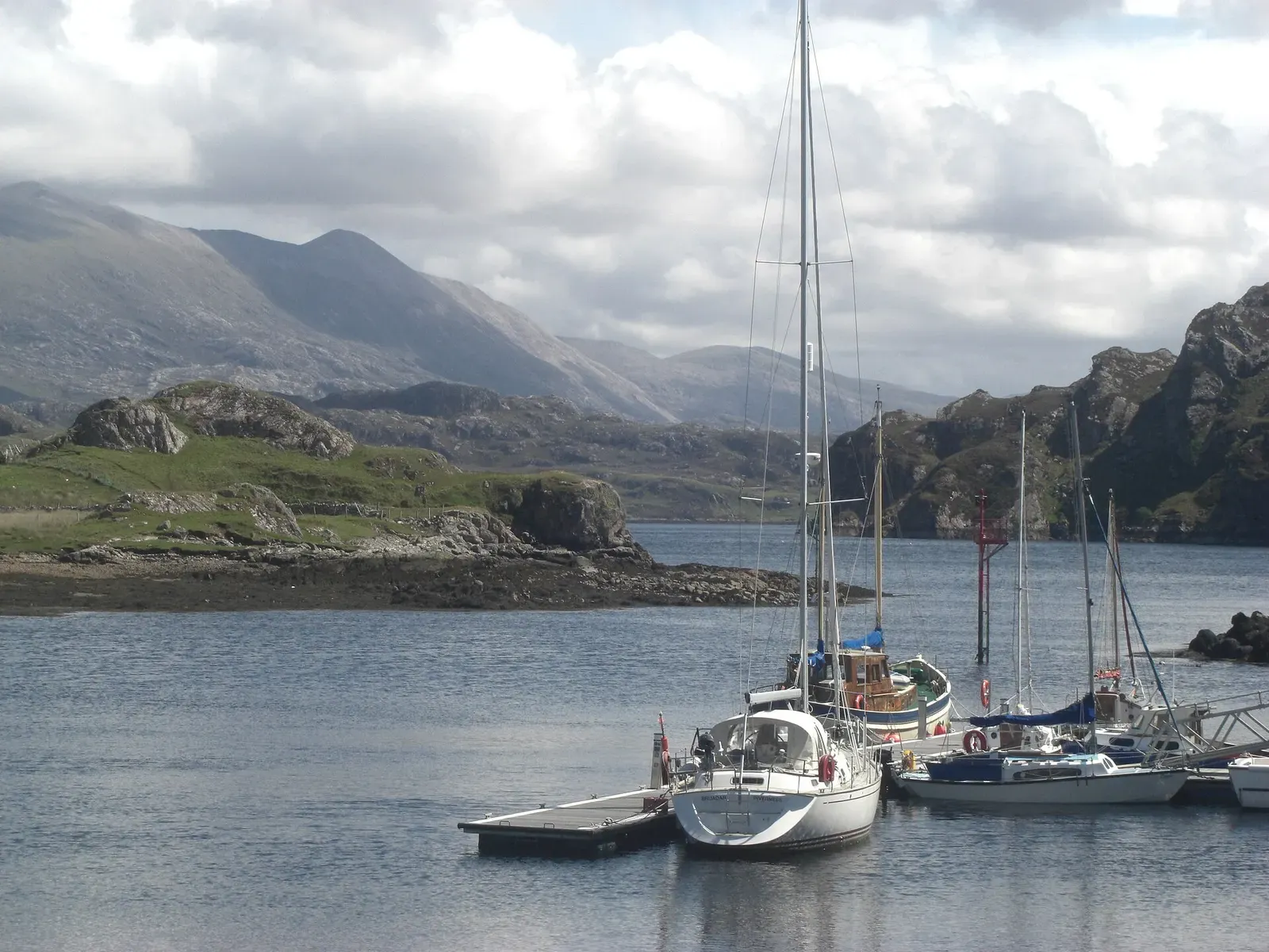 Several white sailing yachts moored at a pontoon on calm grey water, with rocky islets and cloud-capped hills behind — Kinlochbervie & the North-West Highlands