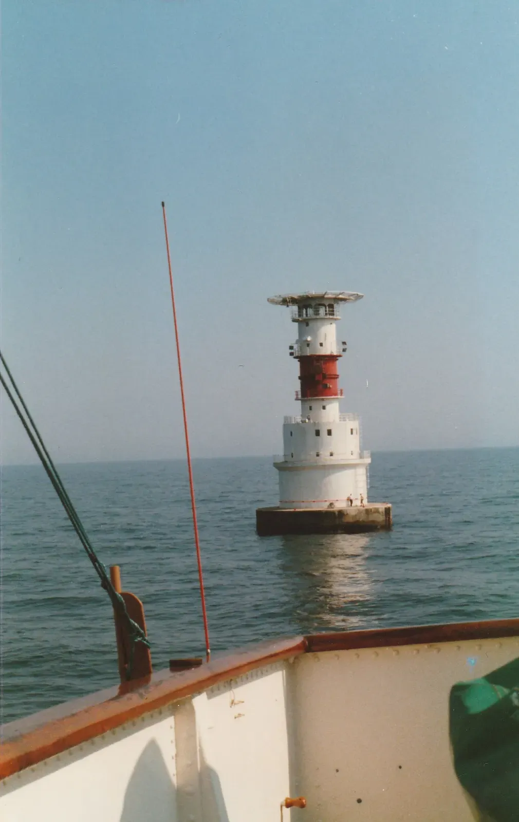 A red and white banded lighthouse standing in calm sea, seen from the bow of a boat on a hazy day — Bolivar, Dublin Bay.