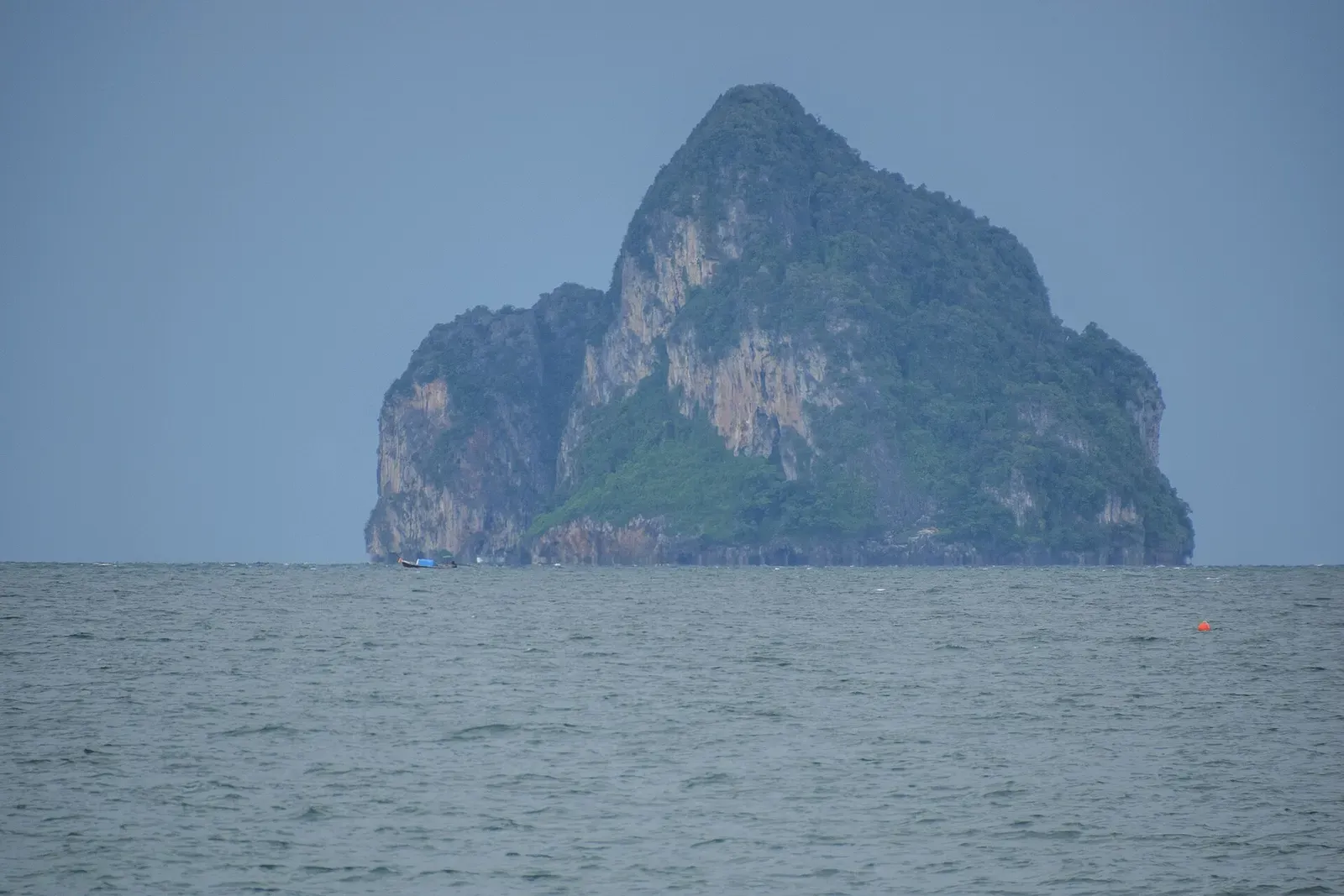 A steep limestone islet with green-topped cliffs rising from a calm grey-blue sea under a hazy sky — Trang Islands