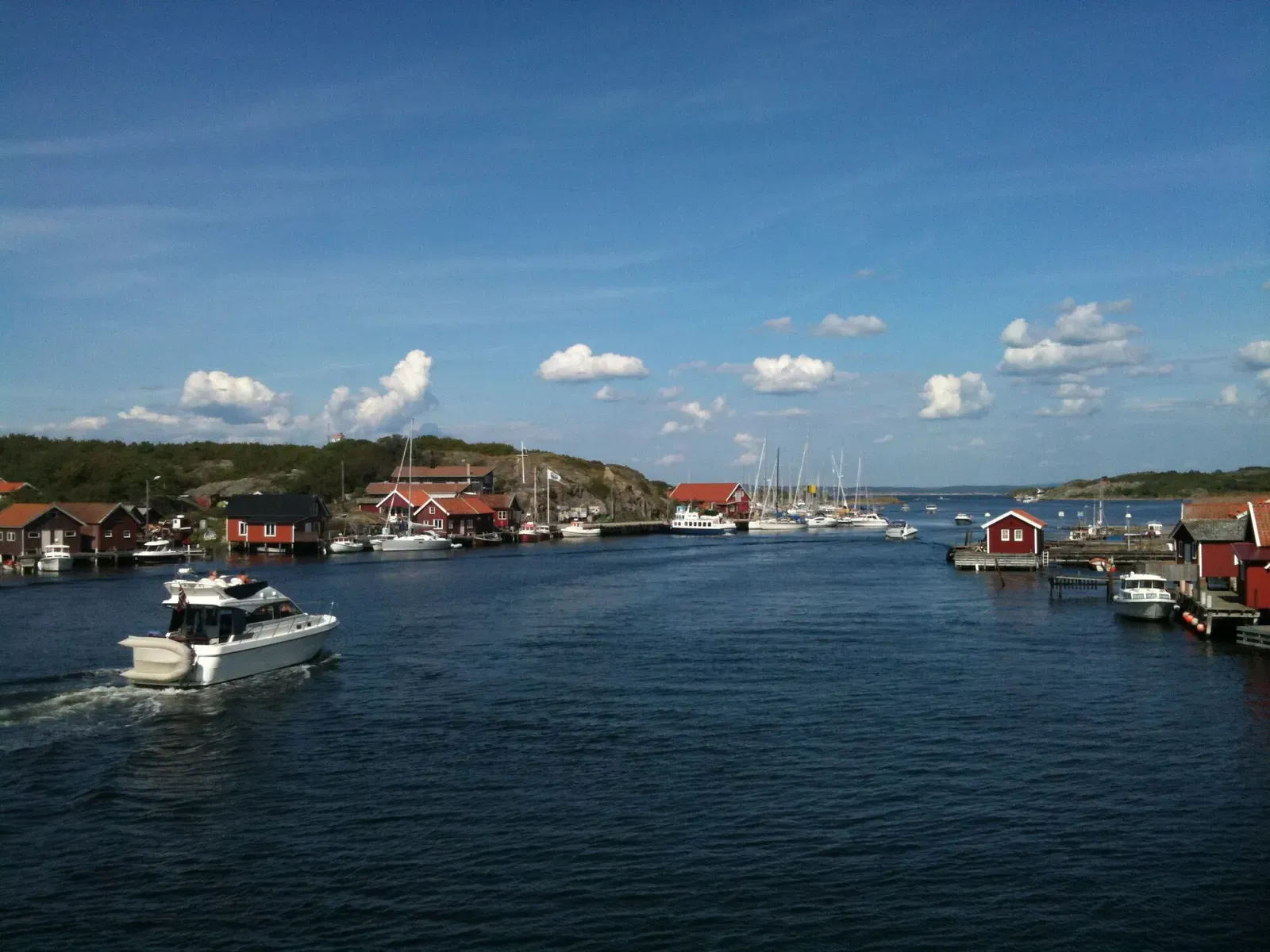 A white motorboat crossing a calm blue sound lined with red boathouses, moored sailing boats and low rocky shores under a blue sky — Koster Islands & Kosterhavet