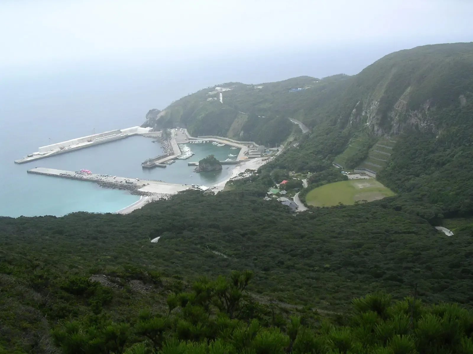 Hazy aerial view of a breakwater harbour and pale blue water below forested green hillsides — Kozushima