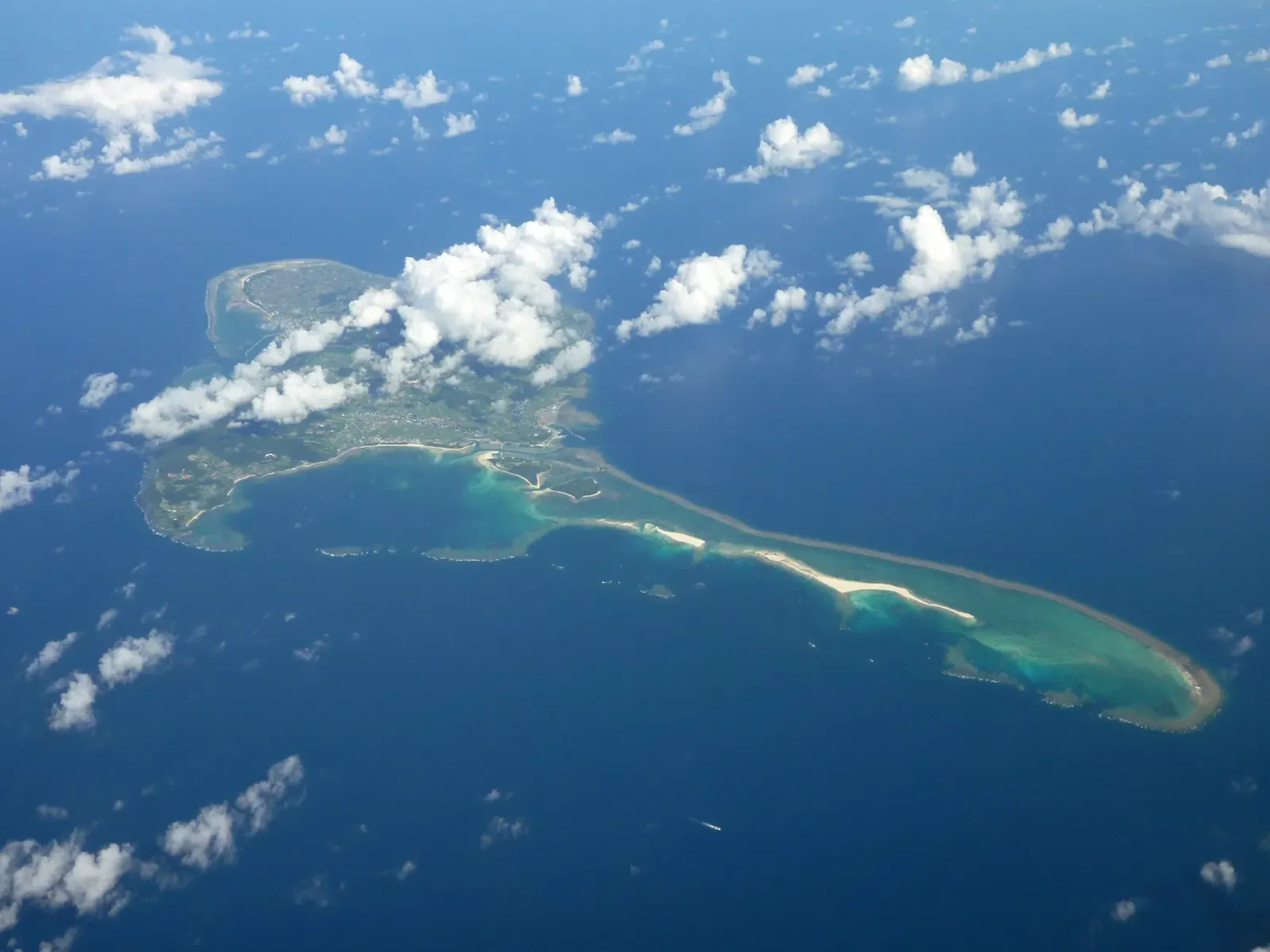 Aerial view of a green island ringed by turquoise reef, with a long white sandbar trailing east into deep blue sea under scattered clouds — Kume Island
