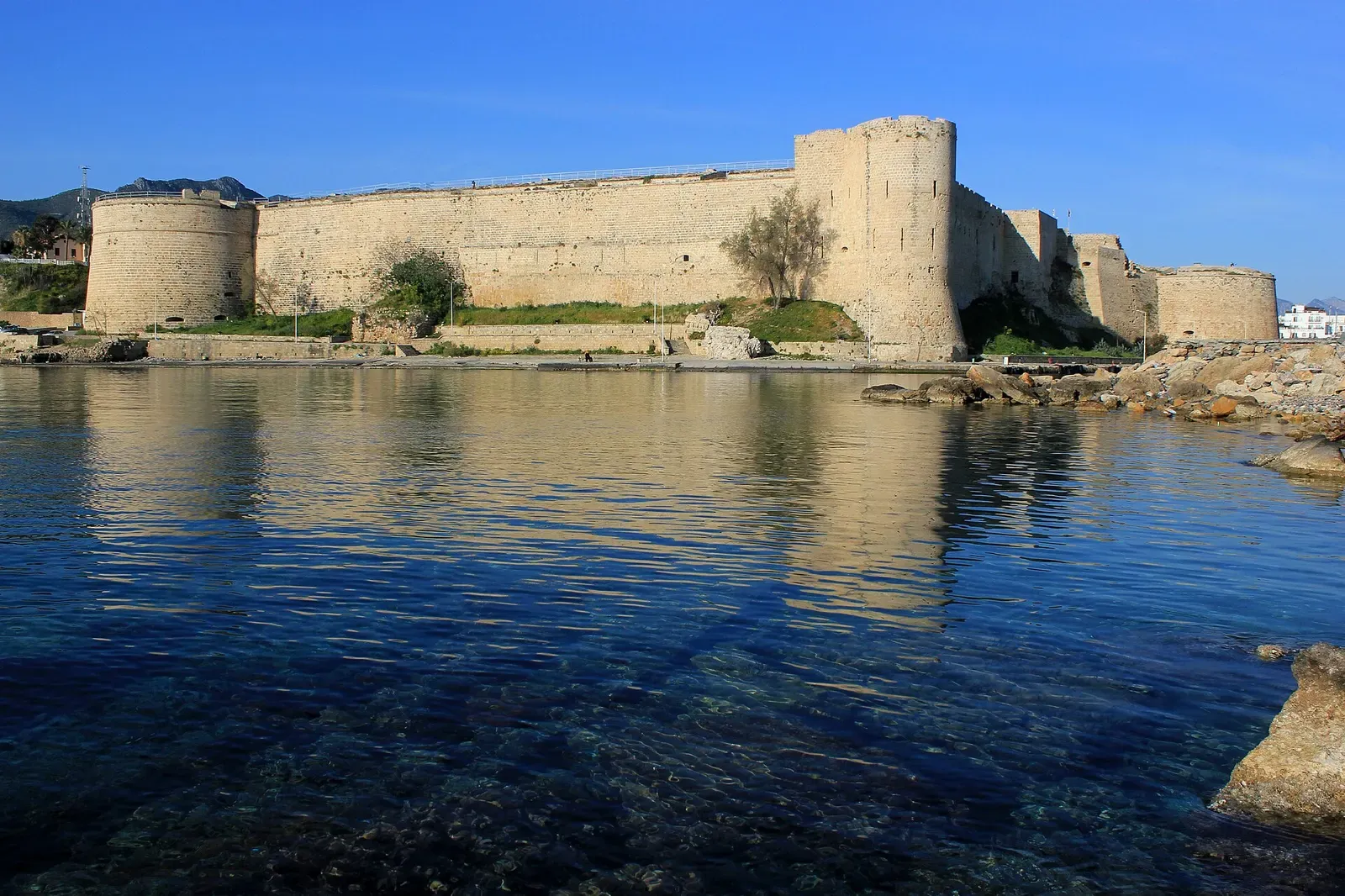 A sand-coloured stone fortress with round towers on a low rocky shore, reflected in calm, clear blue water under a blue sky — Kyrenia (Girne)