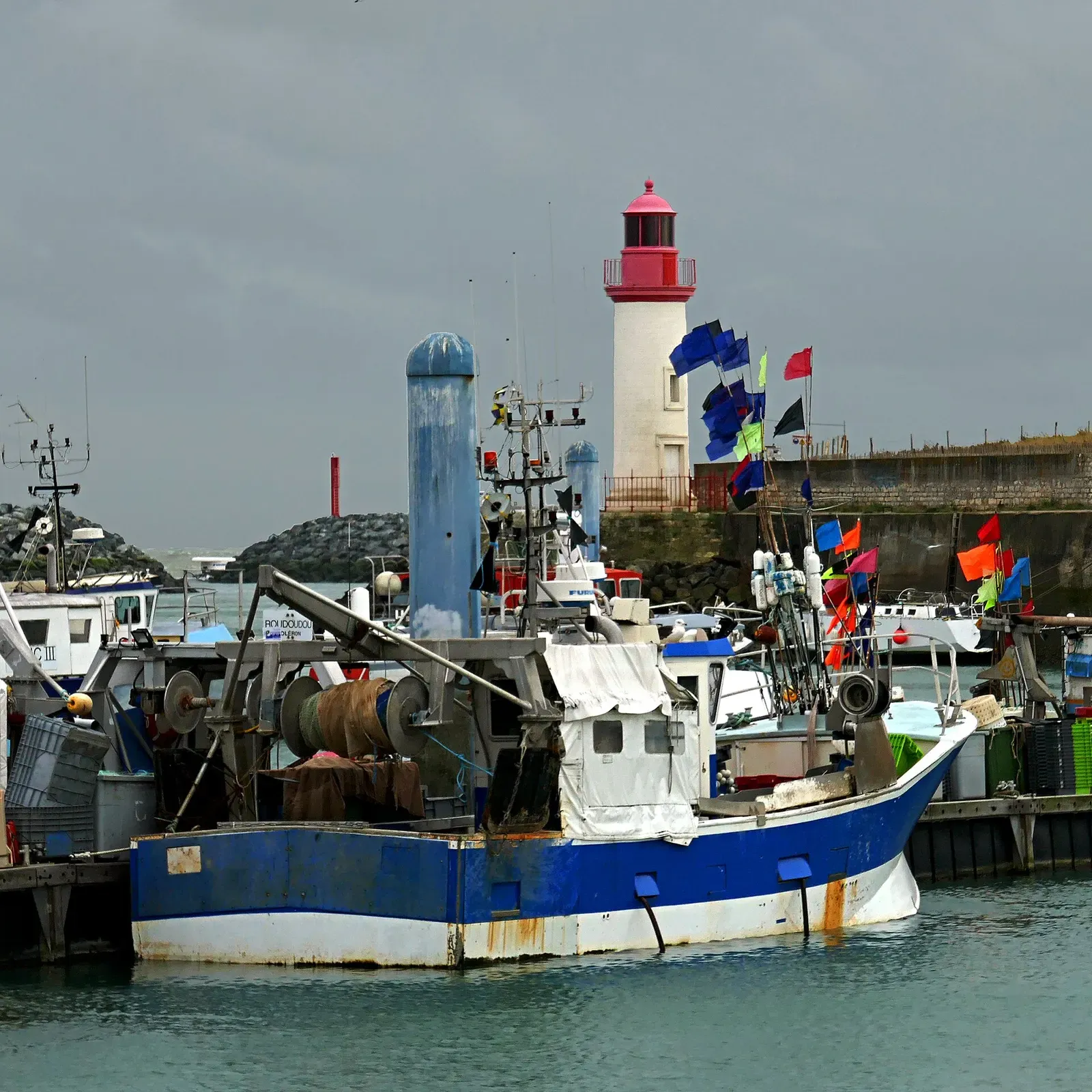 A blue and white fishing trawler moored in a harbour, with a white lighthouse topped in red and colourful flags under a grey sky — Île d'Oléron & the Pertuis d'Antioche