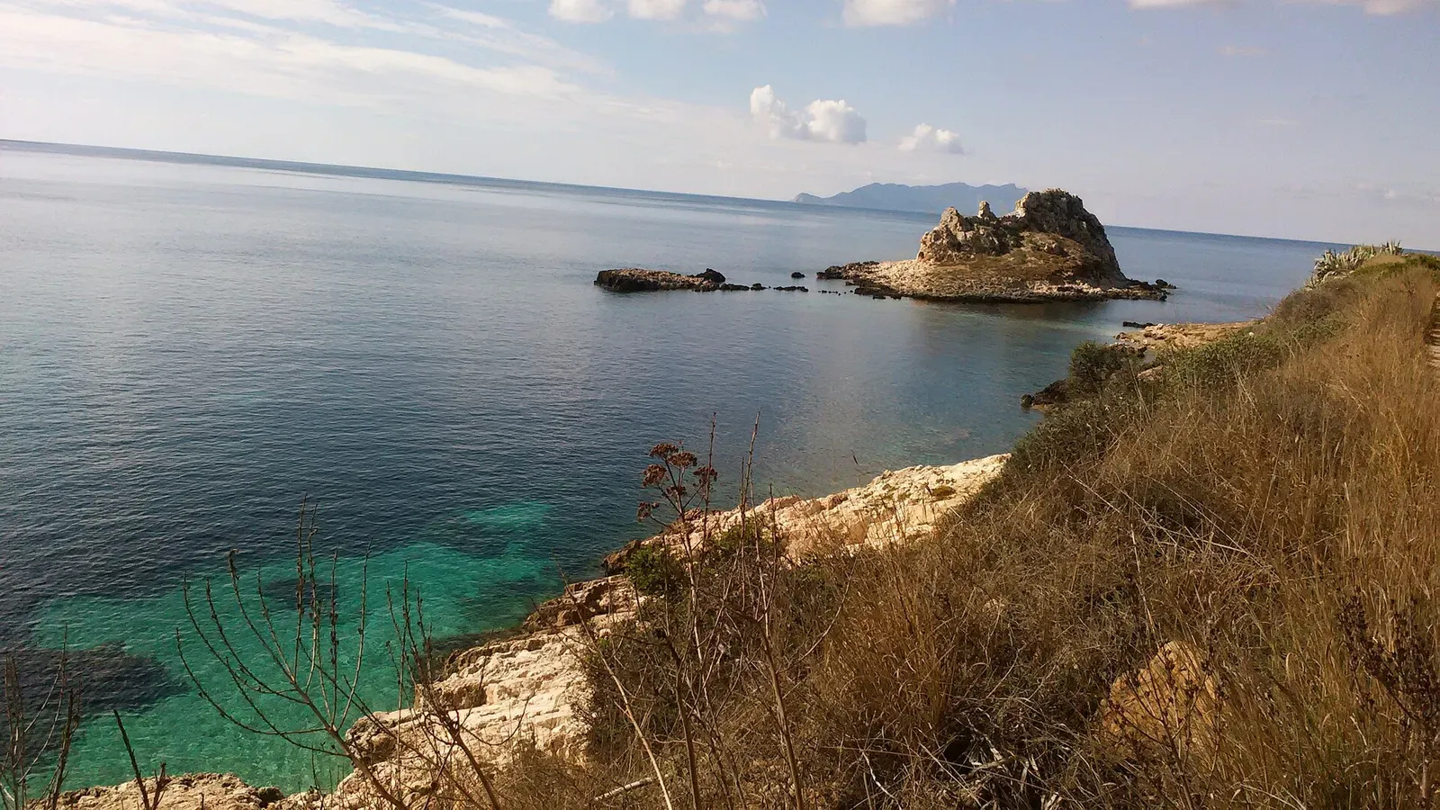 A rocky islet with a jagged crest sits off a scrubby limestone shore, with clear turquoise shallows and a distant island on the horizon — Egadi Islands