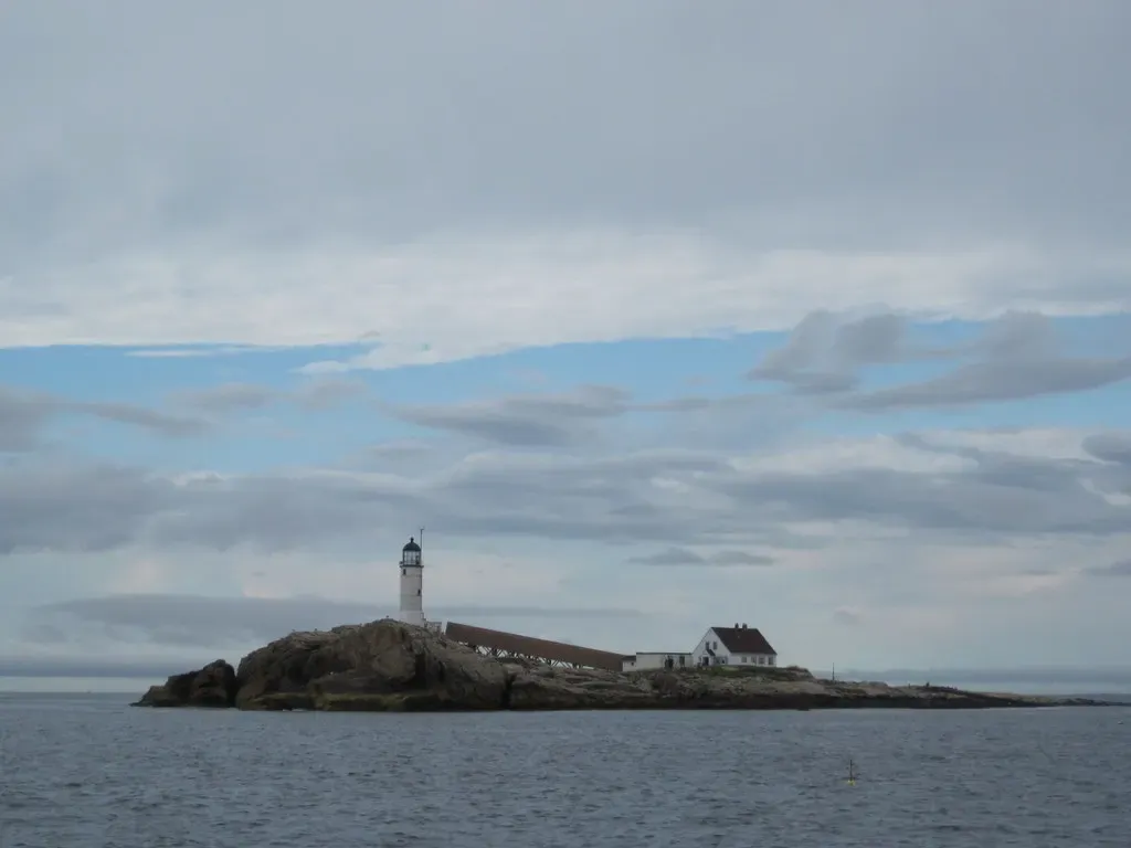 A low rocky islet with a white lighthouse and a long white keeper's building, seen across grey sea under a cloudy sky — Isles of Shoals