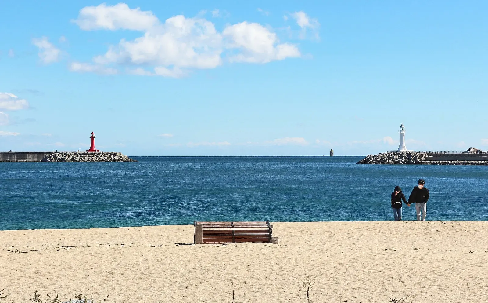 A red and a white lighthouse on rock breakwaters either side of a harbour mouth, seen across blue sea from a sandy beach with two people walking — Sokcho & Goseong.