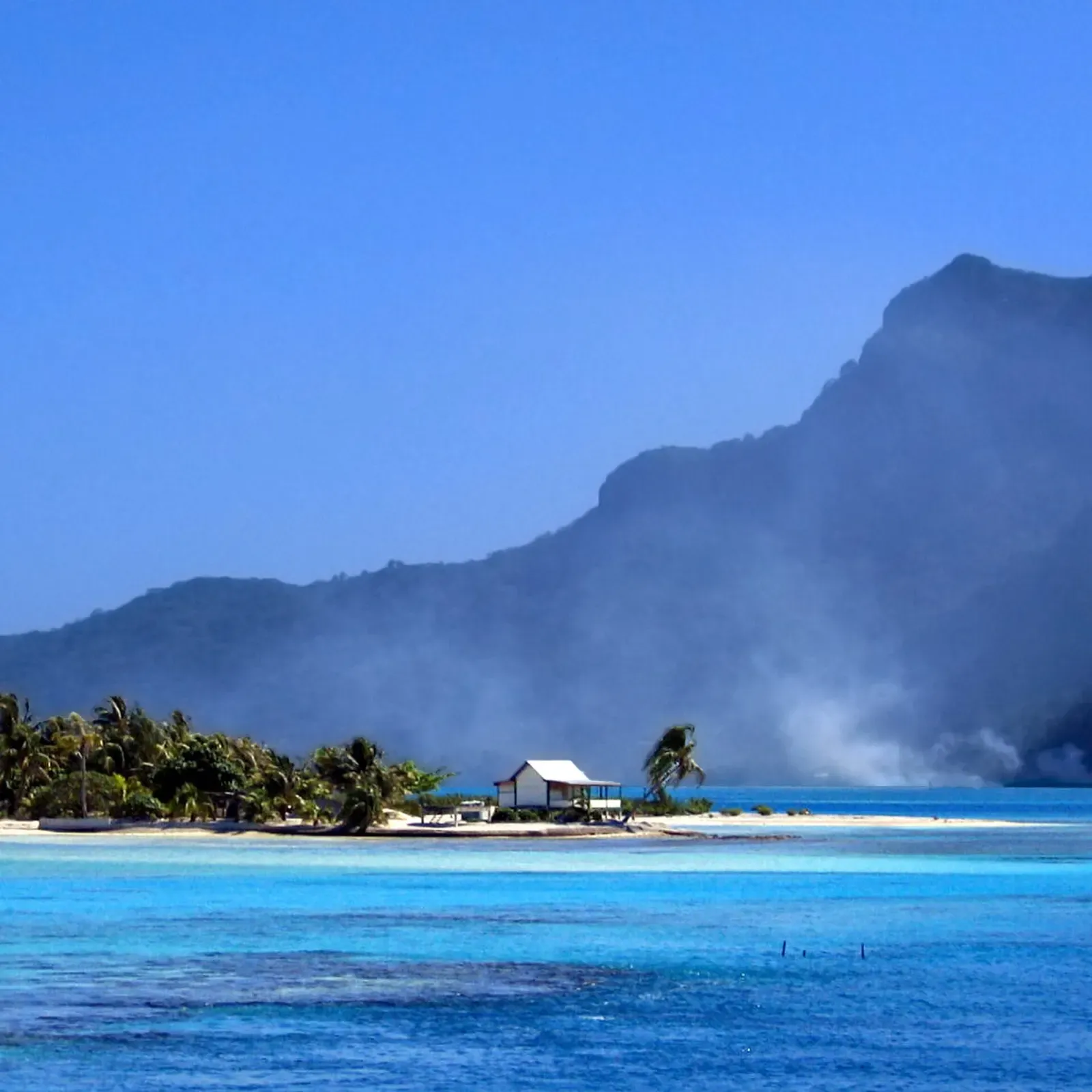A palm-covered motu with a small white house on a sand spit, turquoise lagoon water in front and a hazy green mountain behind — Maupiti.