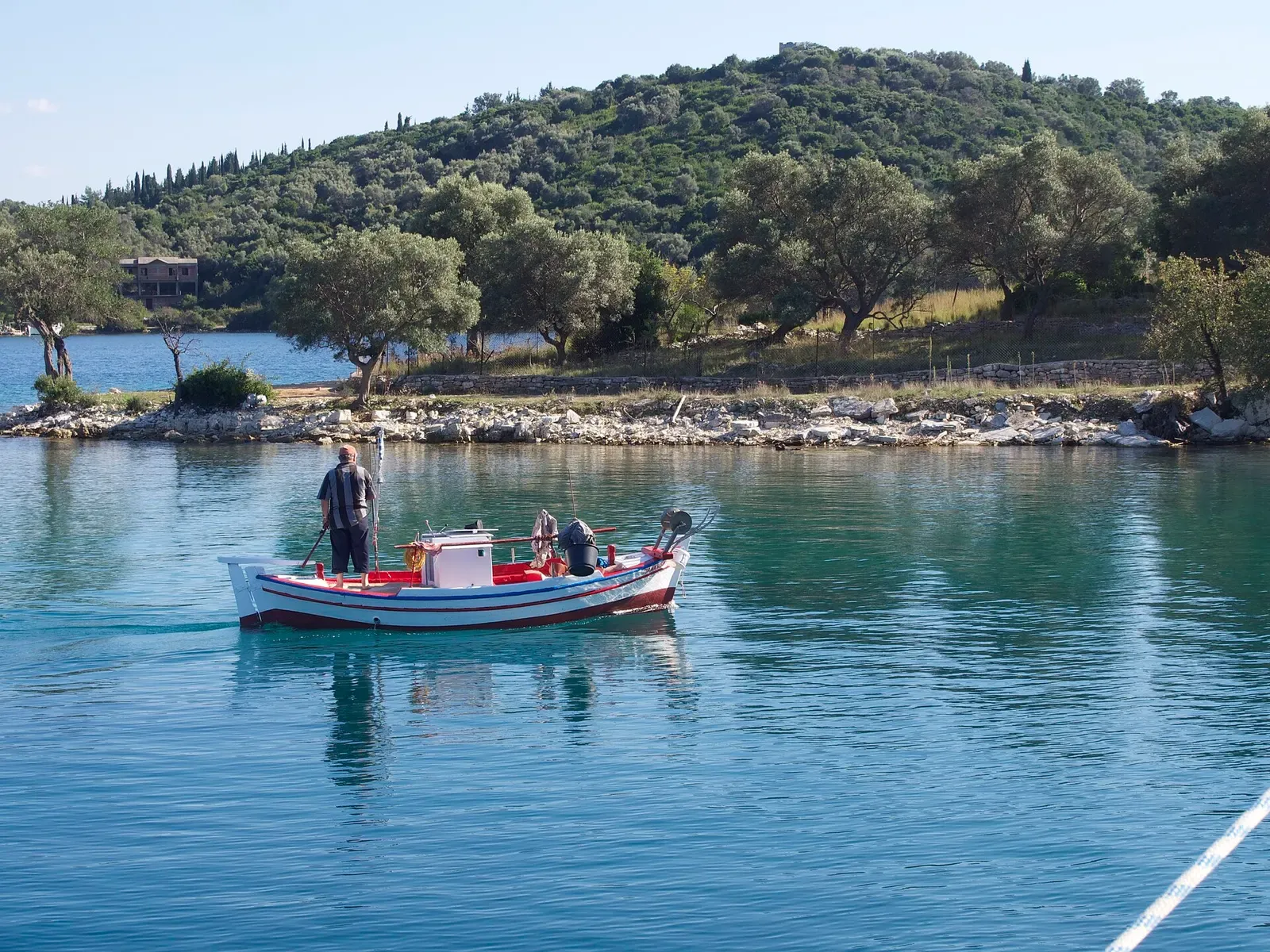 A white wooden fishing boat with red and blue trim crosses calm turquoise water past an olive-covered headland with a rocky shore — Lefkada & Meganisi