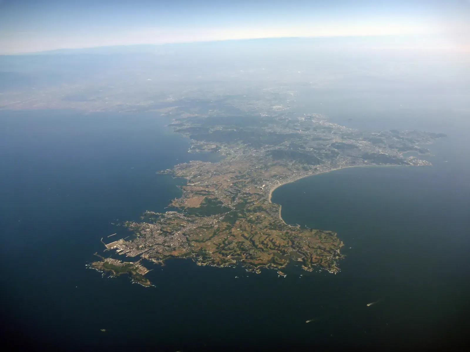 Aerial view of a green, farmed peninsula with a small island at its tip, set in dark blue sea under haze — Miura Peninsula