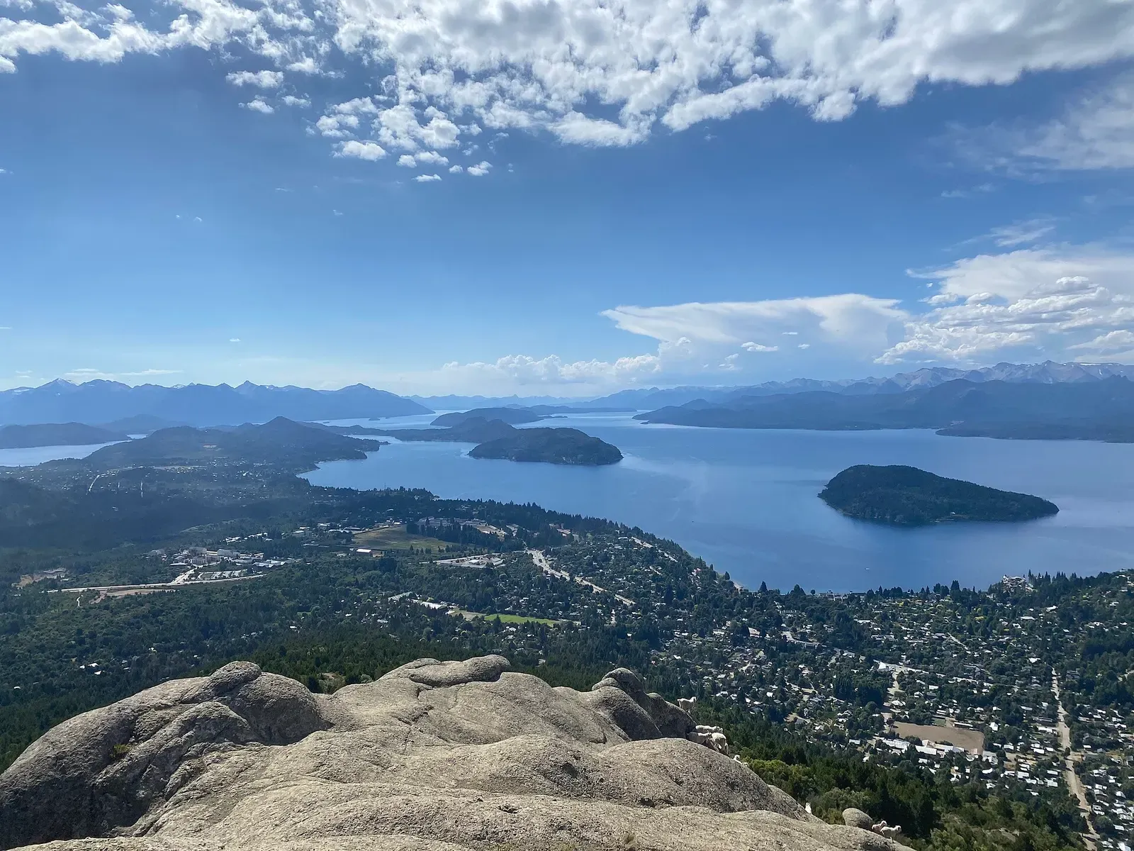 A wide blue lake dotted with wooded islands under scattered cloud, seen from a granite summit above a forested town — Lago Nahuel Huapi & Bariloche