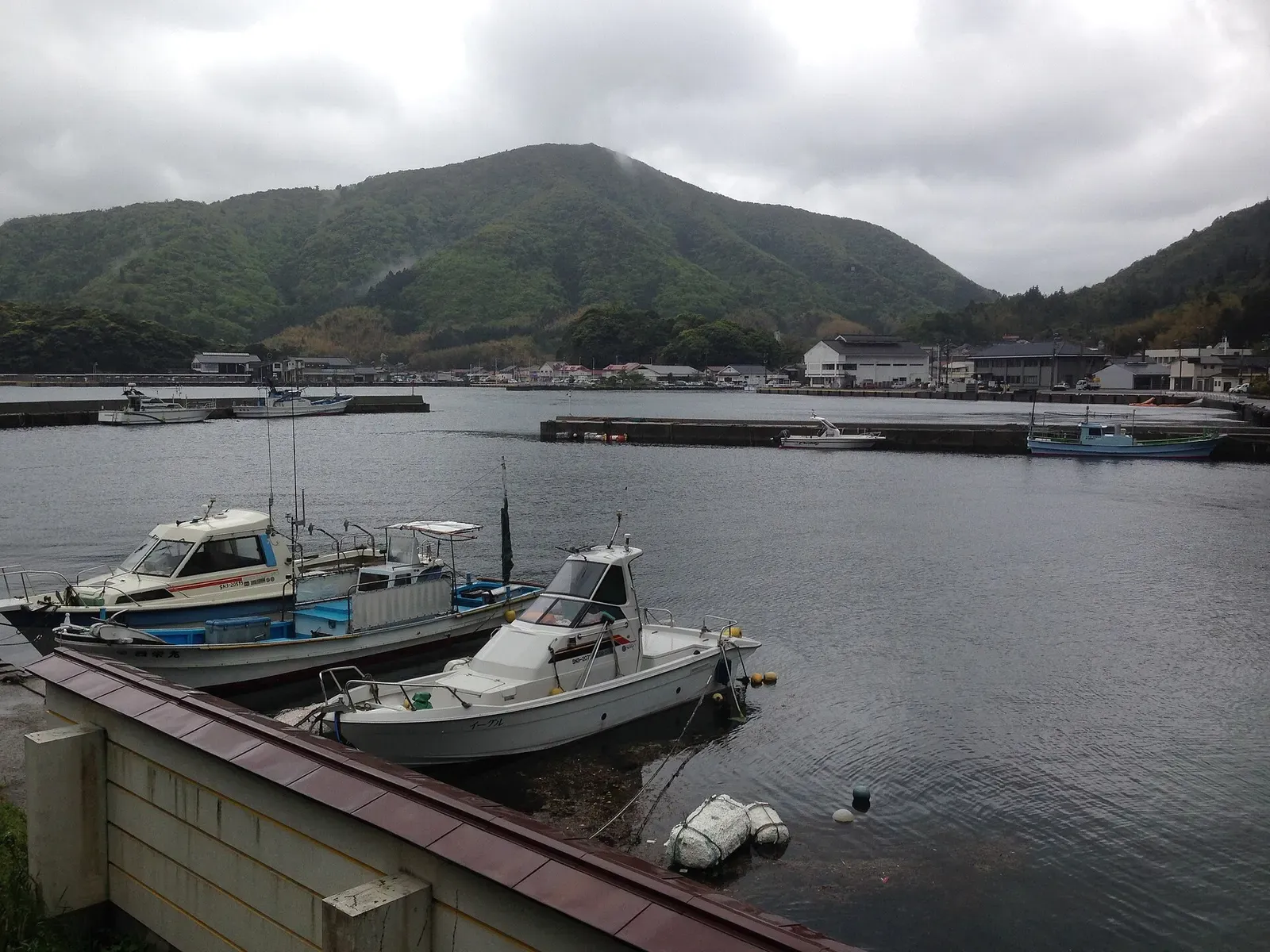 Three small motorboats moored by a harbour wall, with a village and a green forested hill under grey cloud behind — Oki Islands