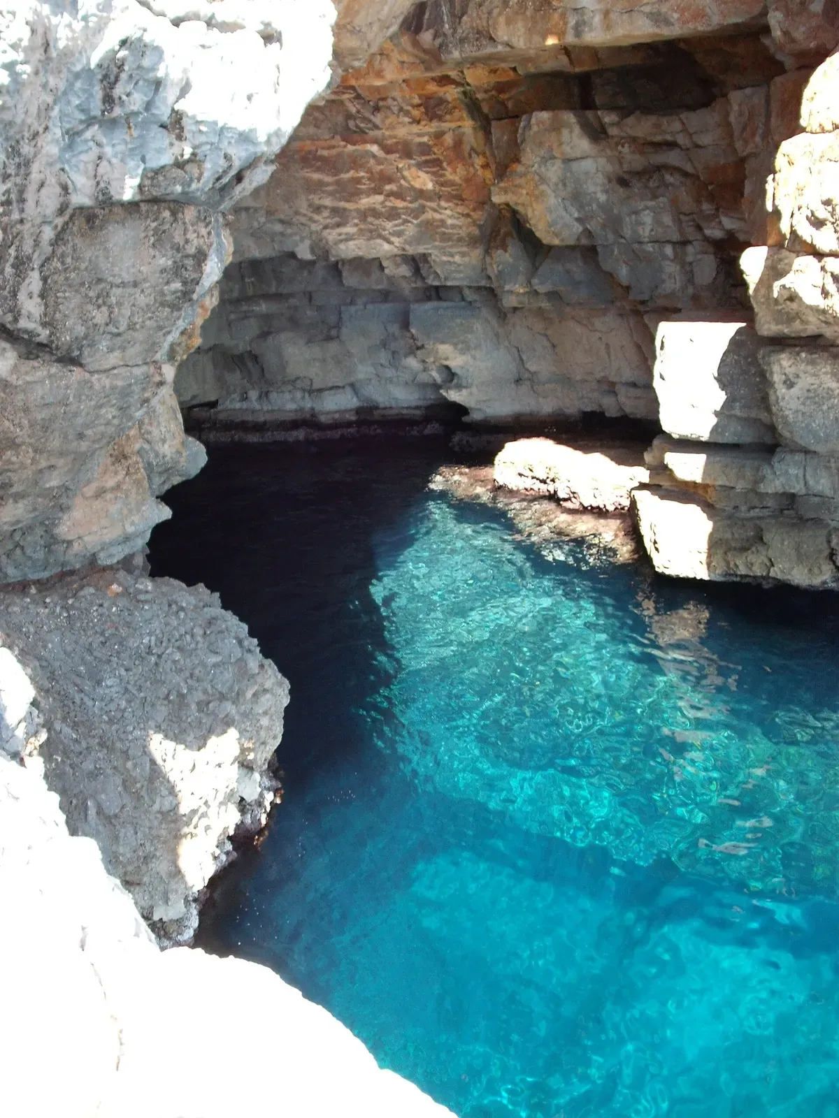 Looking down from a rock ledge into a cave mouth with clear turquoise water over pale rock beneath an overhanging limestone roof — Odysseus' Cave, Mljet National Park