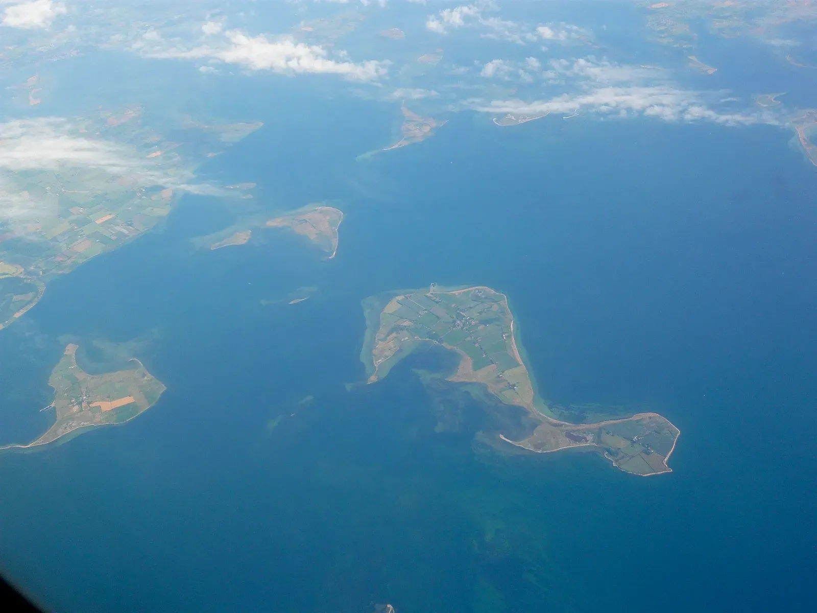 Aerial view of several low green islands with field patterns and pale shallows in a deep blue sea, with thin clouds at the top — South Funen Archipelago