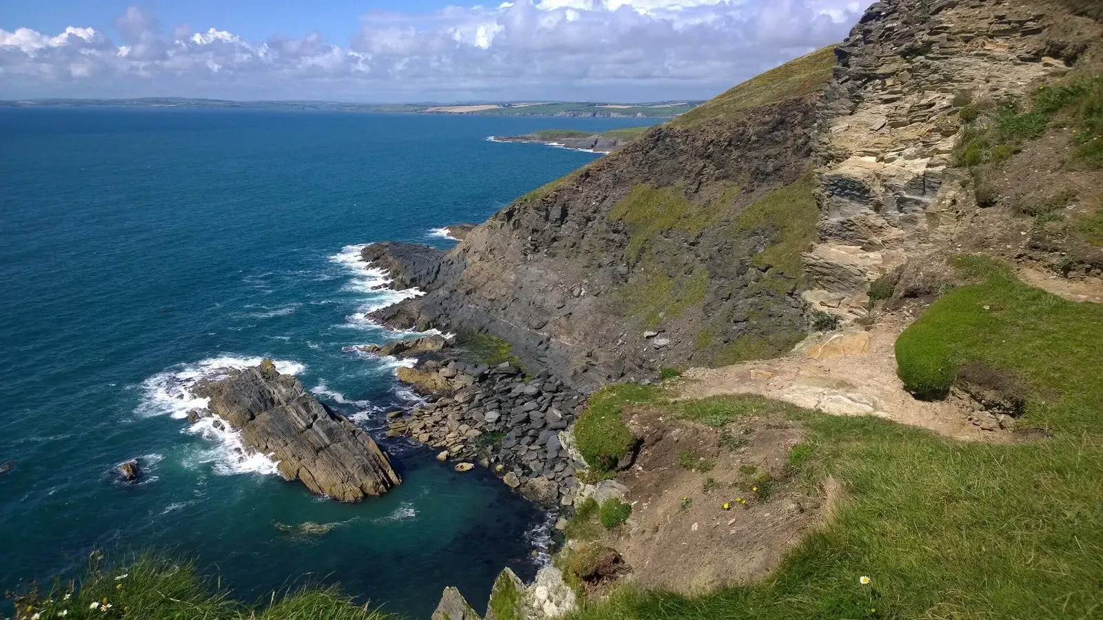 Grassy cliffs drop to rocky ledges and white surf along a blue sea, with a long coastline stretching into the distance — Kinsale & Old Head.