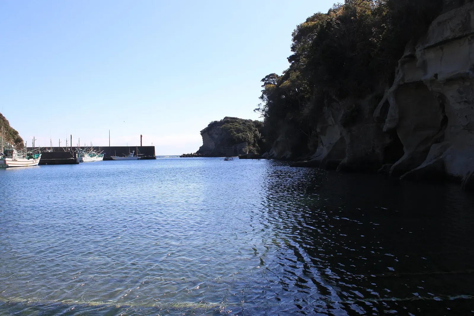 A calm inlet with fishing boats moored at a harbour wall, framed by wooded sandstone cliffs under a clear sky — Katsuura & Namegawa