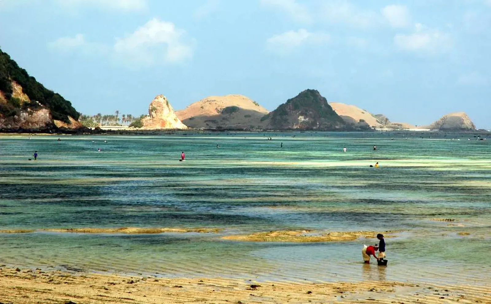 Shallow turquoise bay at low tide with exposed reef flat, two people wading near the sand and dry rocky hills on the far shore — Belongas Bay & South Lombok
