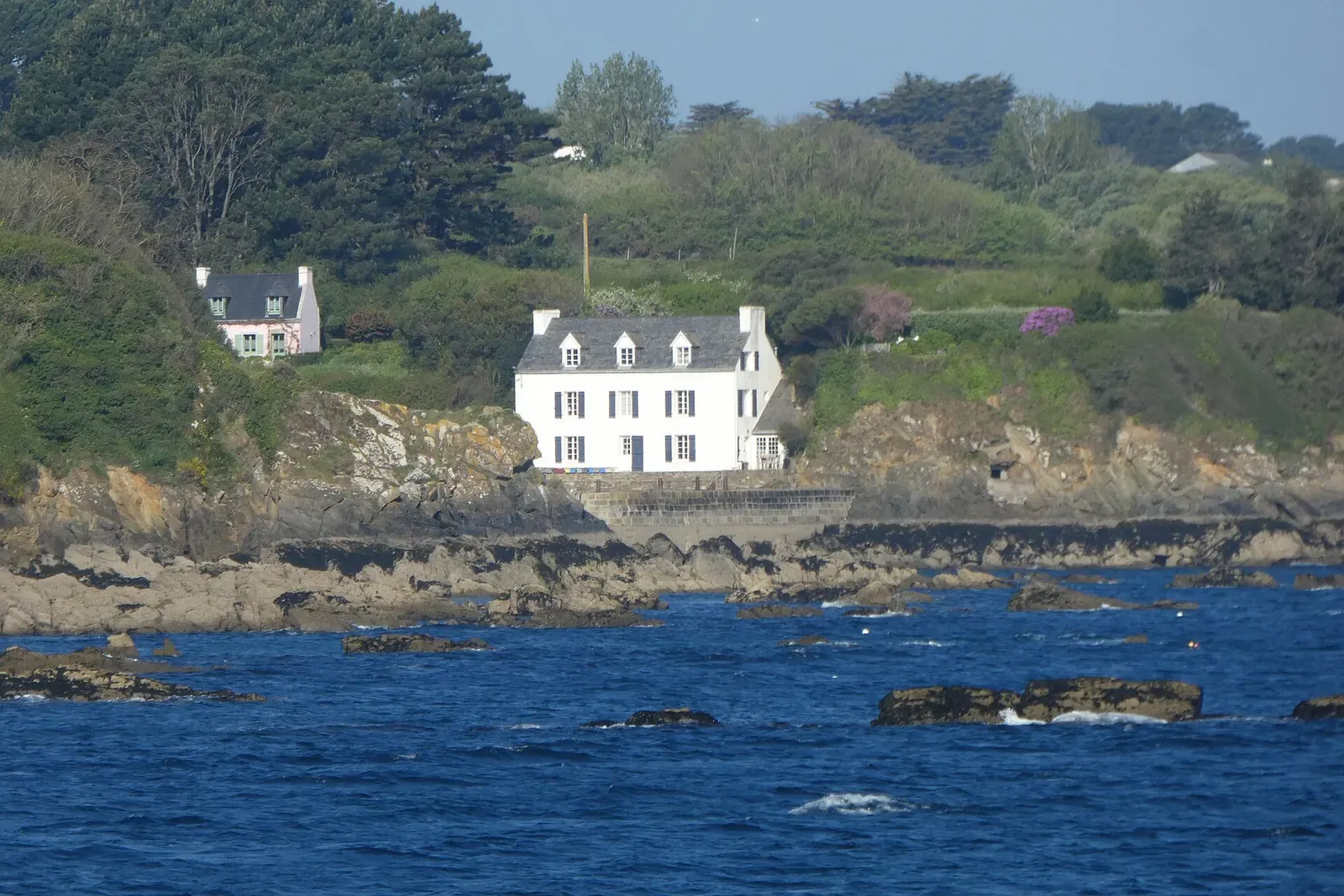 Rocky shoreline with dark reefs in blue water below a white house and green wooded slopes — Île de Groix & Lorient